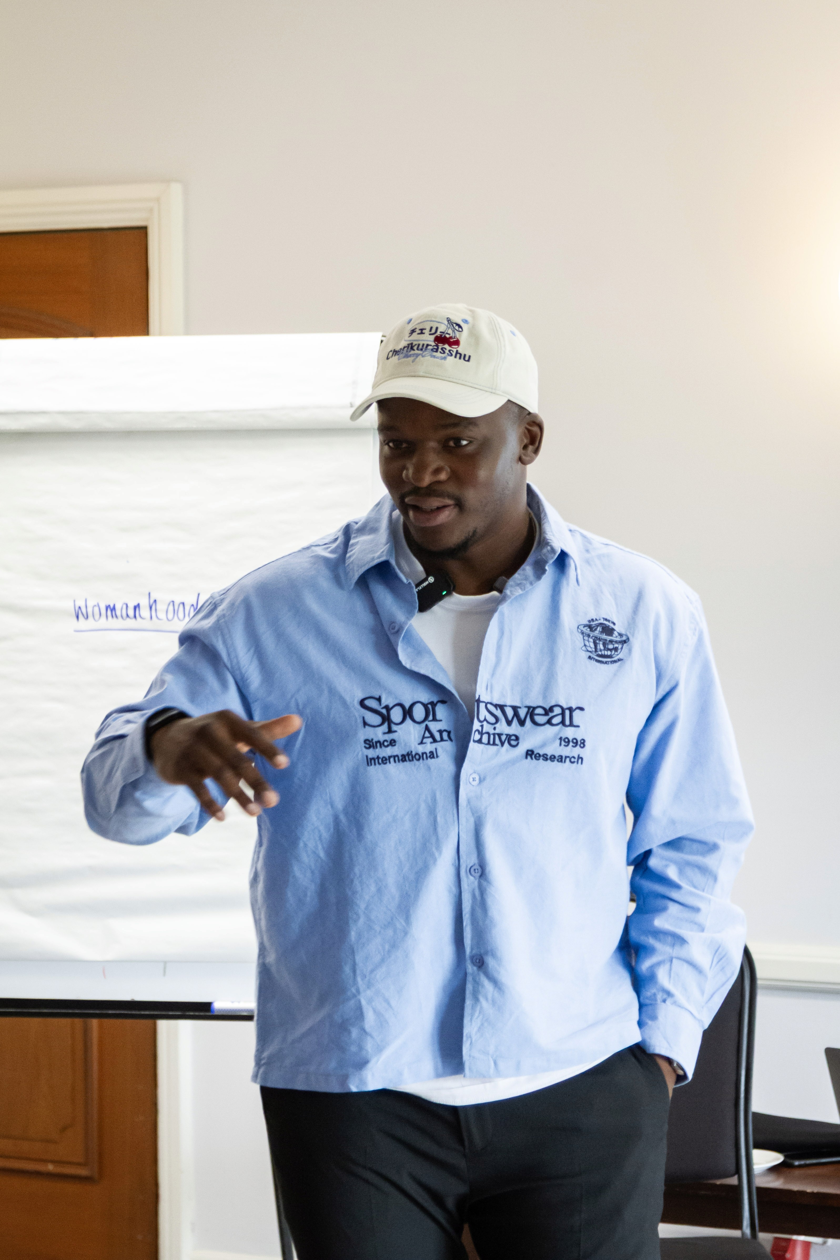 A person wearing a light blue jacket and cap gestures while speaking in front of a presentation board.