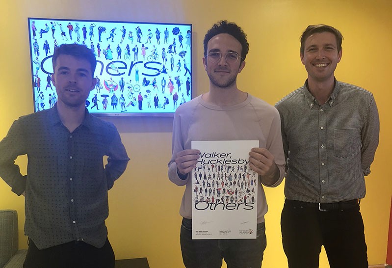 A photo showing three people sitting in an exhibition room. One of them is holding a posters with human drawn peoples and the text: Others. Also behind them there is a tv screen with the same drawings and text.