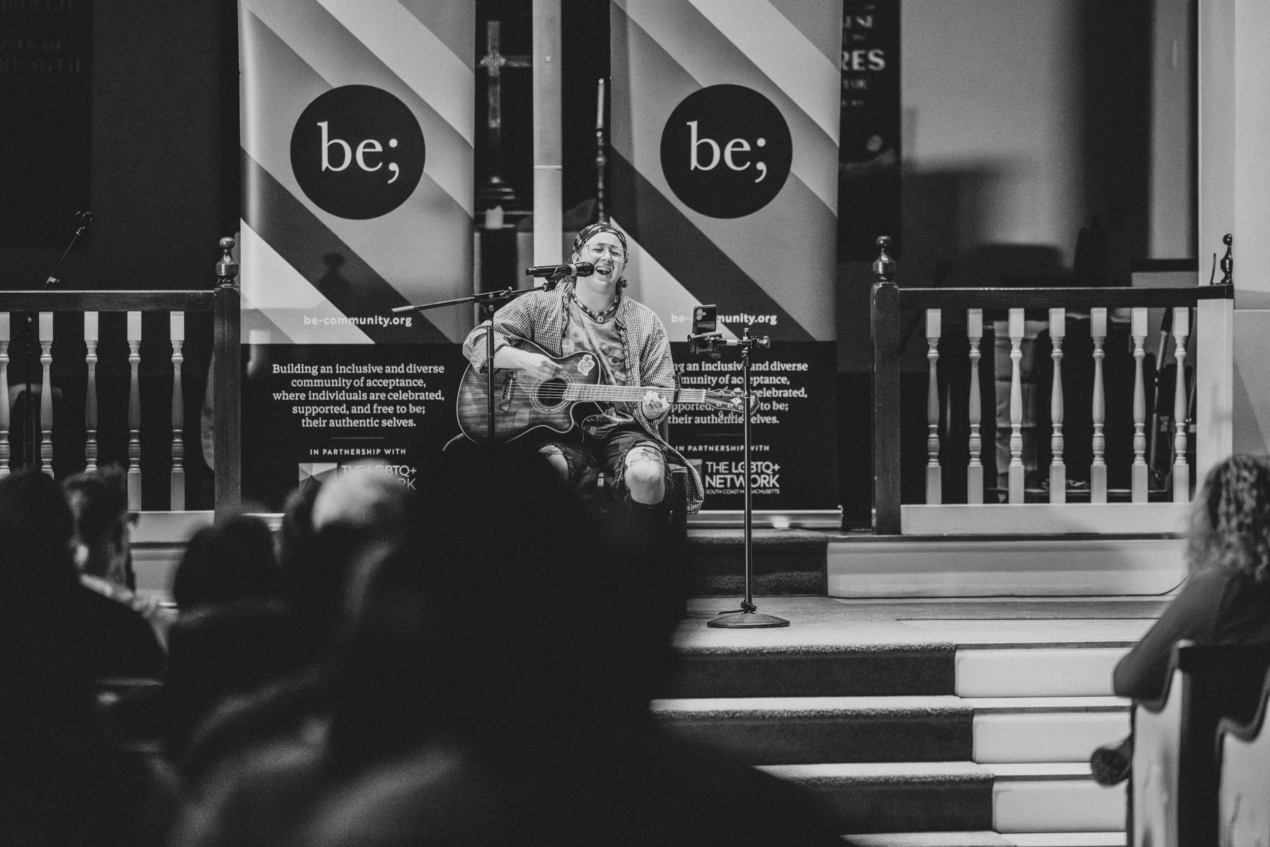 Performer playing guitar and singing on stage at the Queer Open Mic (QOM) hosted by be; community in Bridgewater, Massachusetts