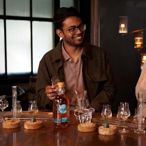 A man is smiling while holding a whiskey glass at a table with various glassware and a bottle of Highland Park Single Malt Scotch Whisky.