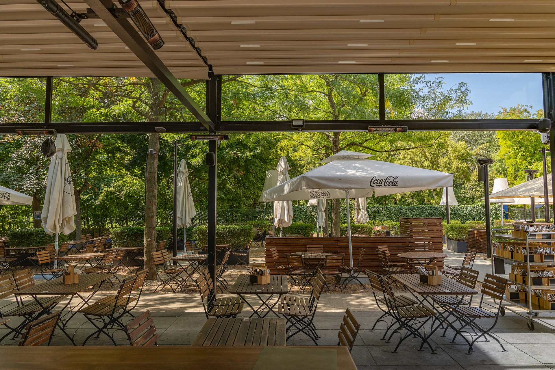 Überdachter Gastgartenbereich mit Holztischen und Blick auf den grünen Außenbereich mit Bäumen.