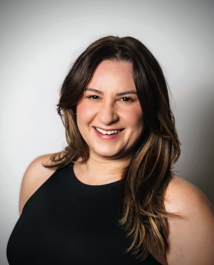 Professional headshot of Krysten Loiselle, wearing a cream blazer over a brown top, standing outdoors with soft sunlight and water in the background, smiling confidently at the camera.