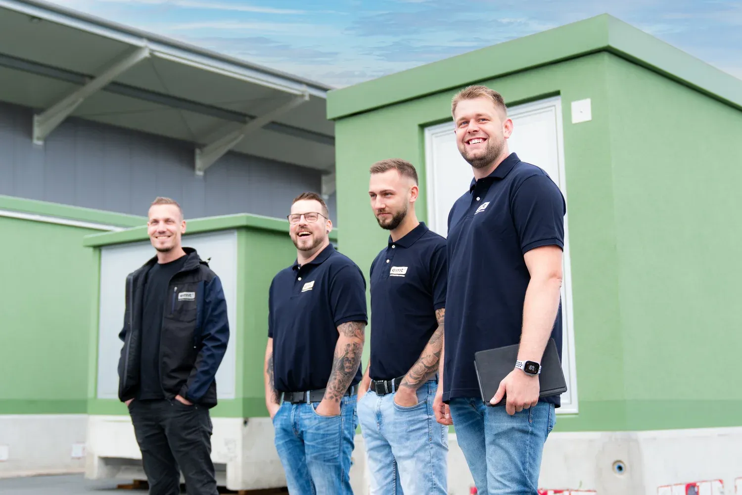 Vier lächelnde Männer in Arbeitskleidung stehen vor grünen Gebäuden.