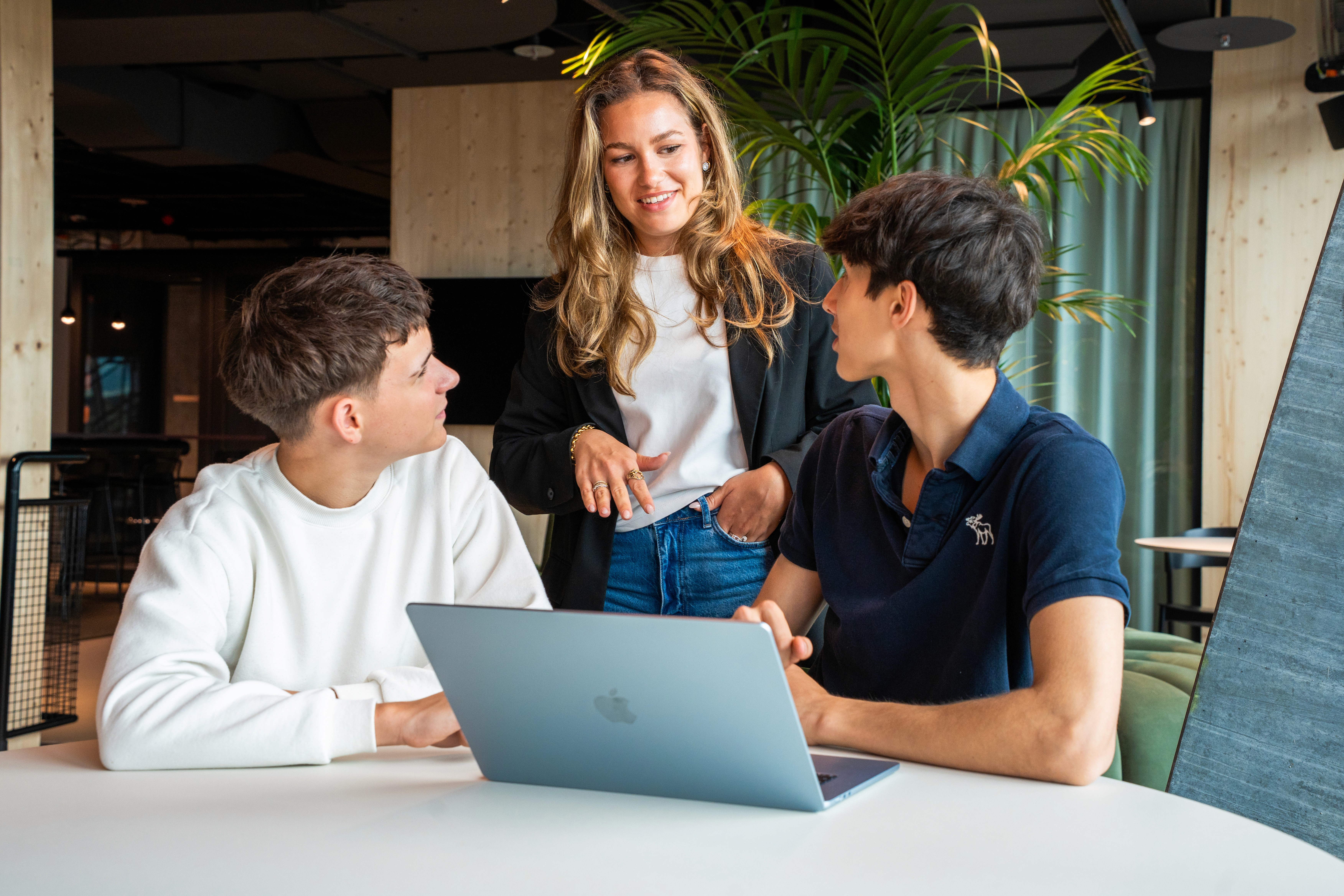 A young person with curly hair smiles at the camera while sitting in front of a laptop against a dark background.