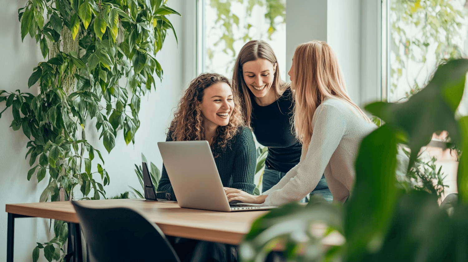 Three women smiling and collaborating around a laptop at a table in a bright office, with plants in the background.