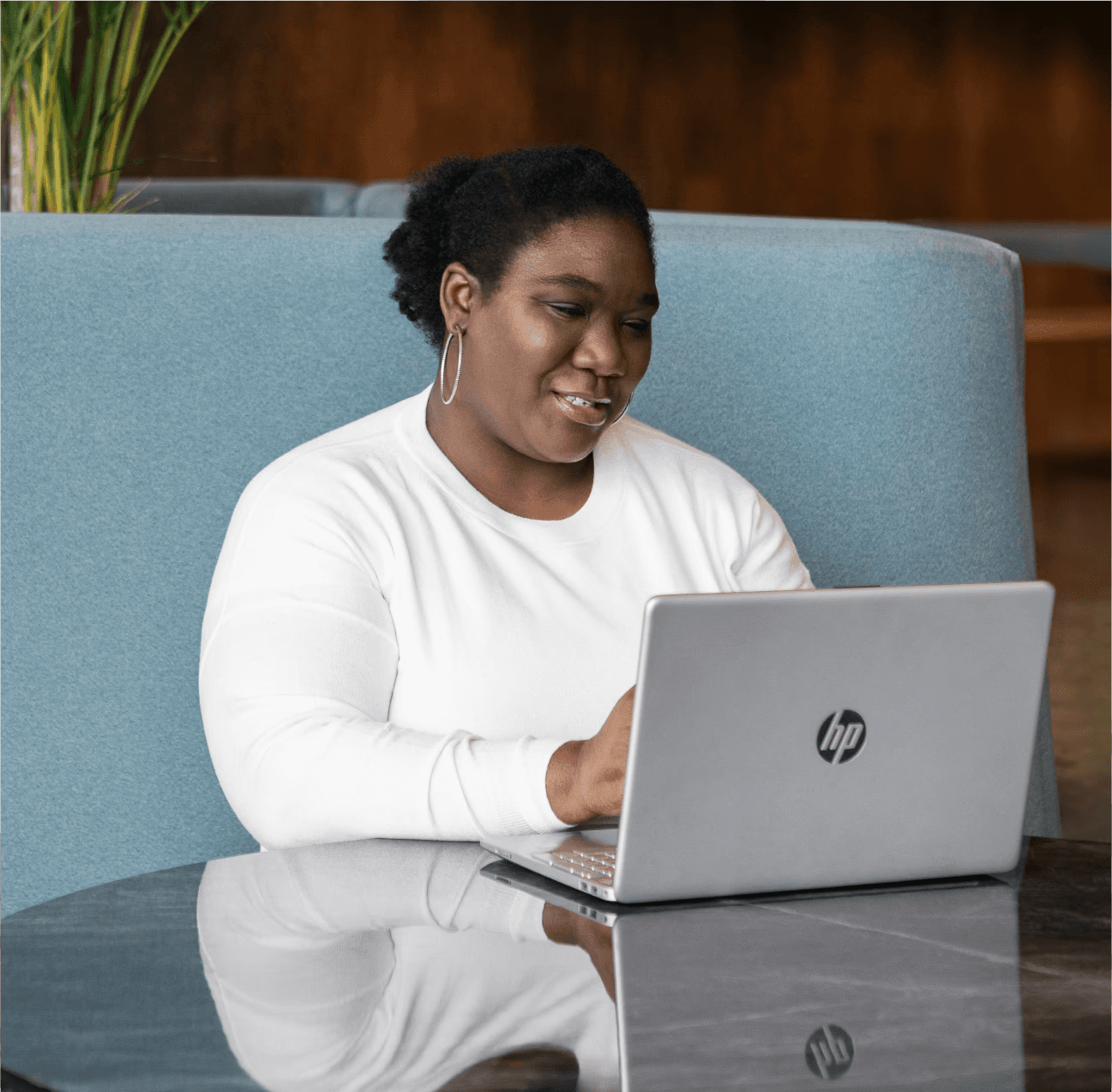 Therapist Sabrina Simpson with her hair pulled back, wearing a white long-sleeved shirt and large hoop earrings. She is seated at a round, reflective dark table, looking down and smiling at an open silver laptop. The background features a plush, light blue sofa and a hint of greenery from a plant on the left. The lighting is soft and natural, highlighting Sabrina Simpson's face and the laptop.