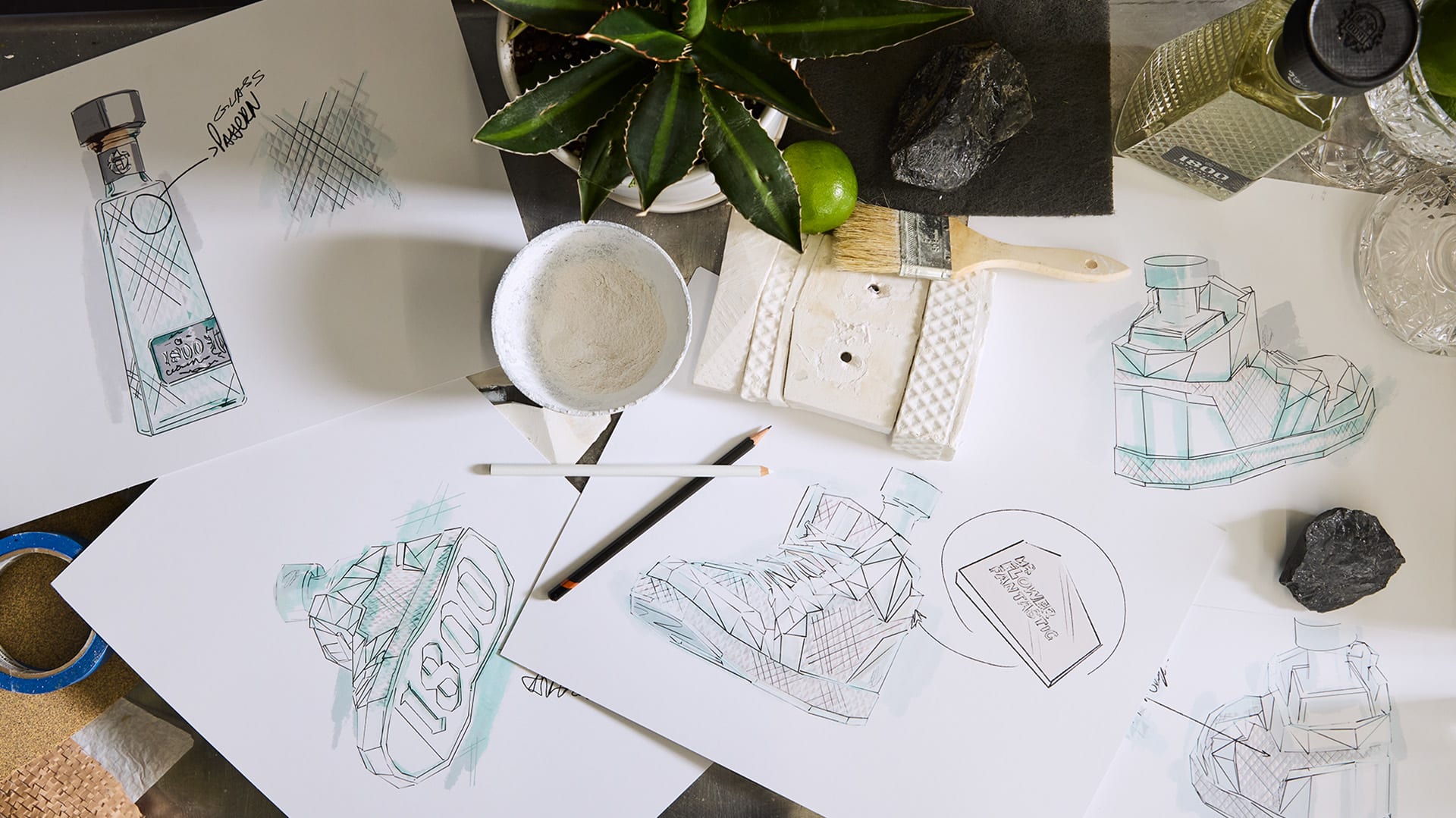 A tabletop filled with design sketches of a glass sneaker bottle, paintbrushes, a mold, a bottle of 1800 Cristalino tequila, and a potted plant