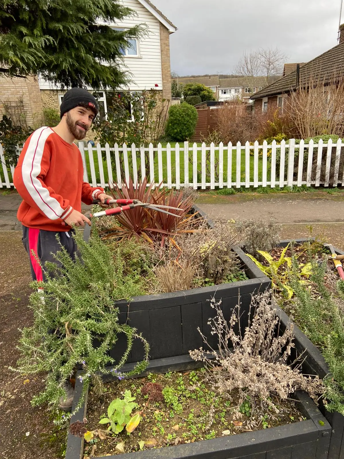 A person in a red jacket tends to a garden bed, surrounded by greenery and a wheelbarrow in the background.