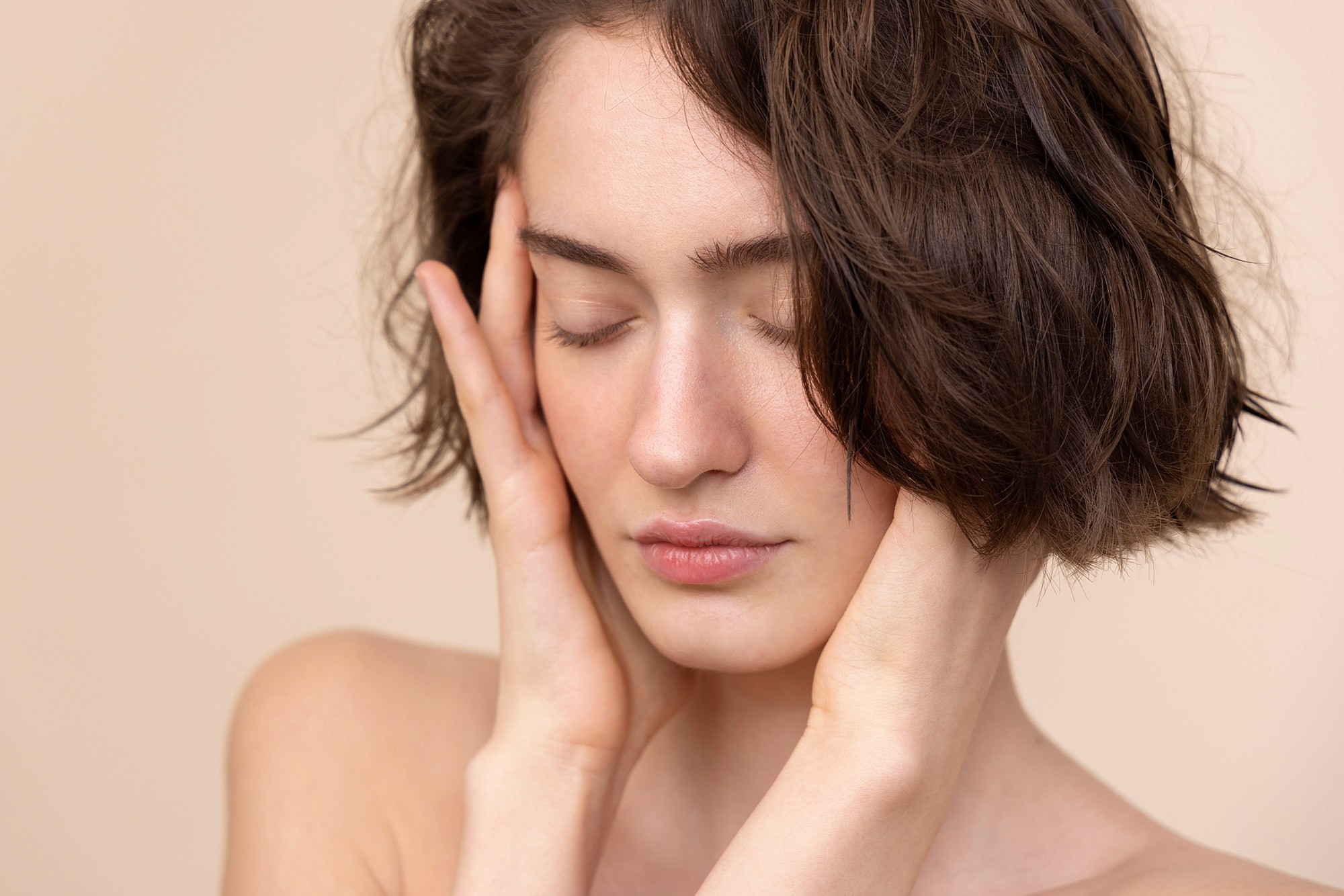 Portrait of a woman with short brown hair, eyes closed, gently touching her face with both hands against a beige background