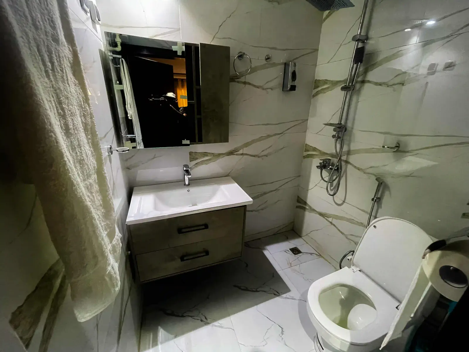 Salle de bain carrelée de blanc avec douche, meuble vasque, miroir et toilettes.