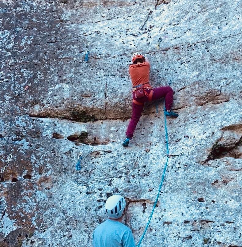 A climber in a red jacket and helmet ascending a steep grey rock face on a rope.