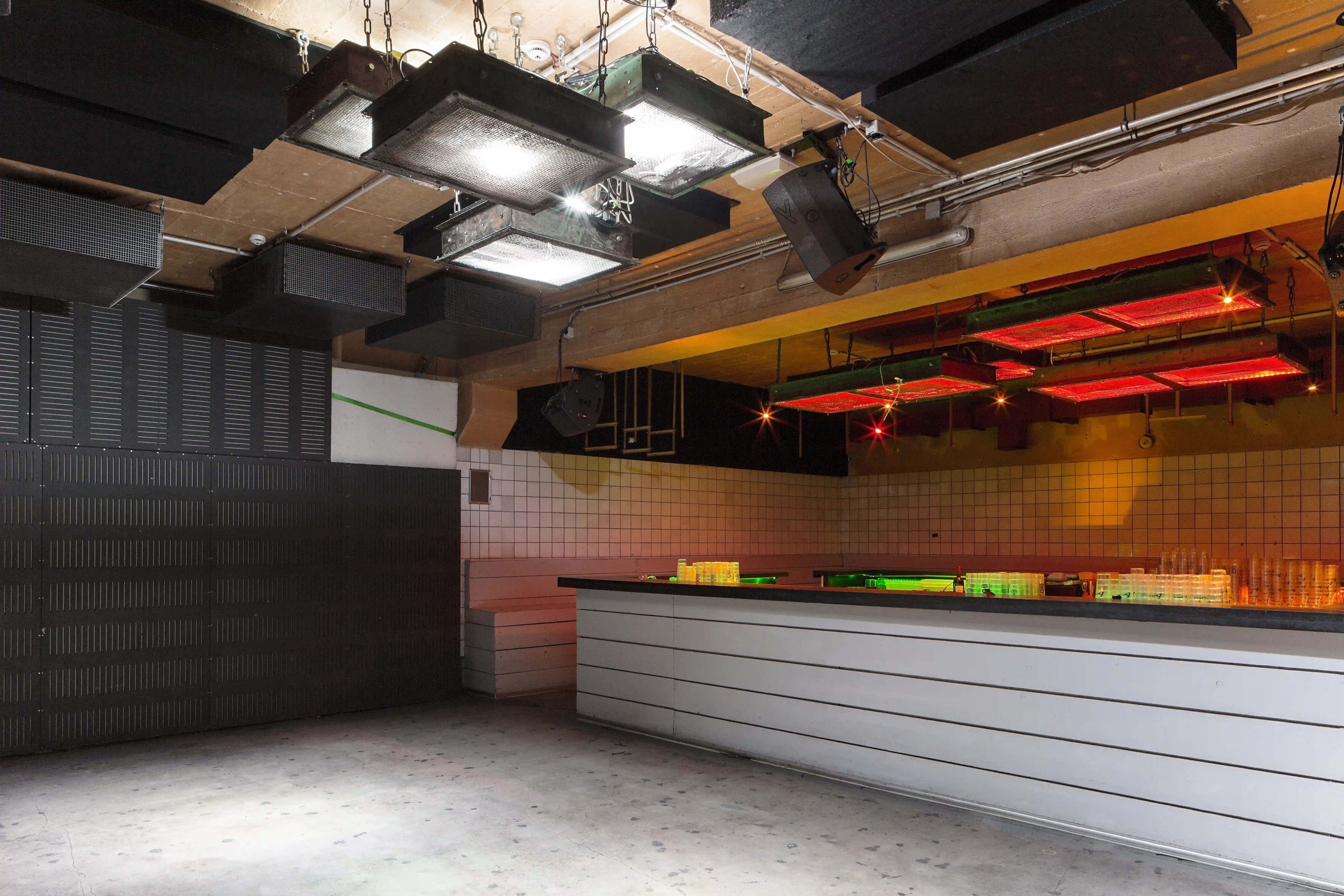 Club room at OHM CLUB with black industrial light boxes hanging from a wooden ceiling, a long white-panelled bar with black top and red-lit grid lights above.