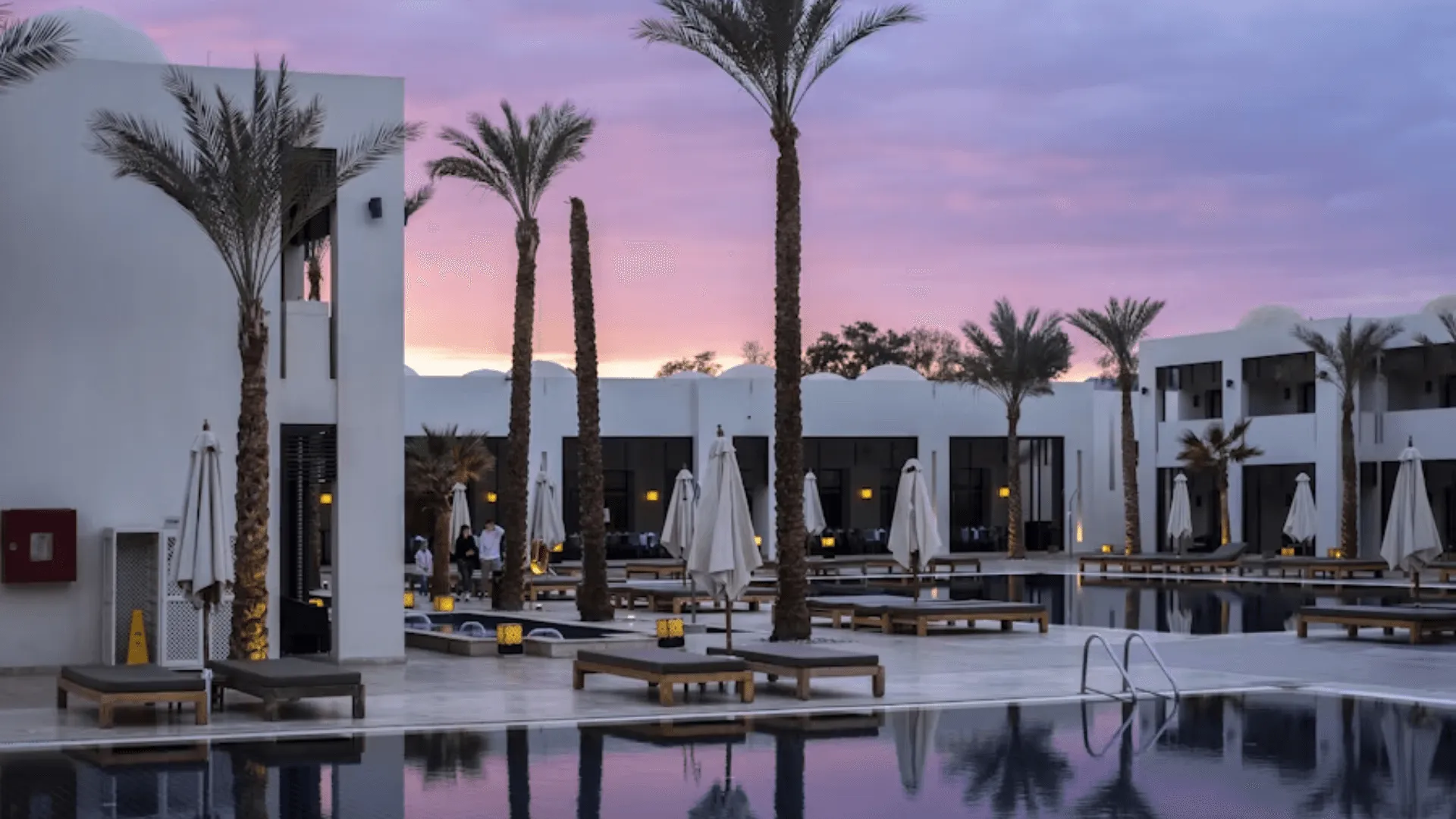 An image of a hotel pool surrounded by palm trees at sunset
