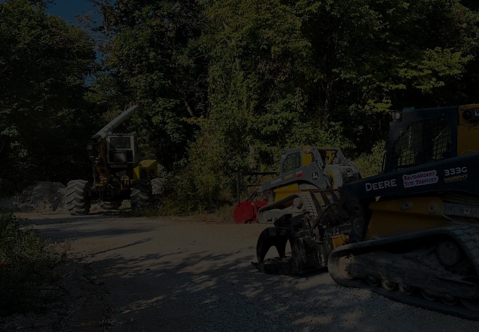 A yellow, tracked vehicle with a large, articulated arm and a bucket at the end. The vehicle is parked on a dirt road in a grassy field with trees in the background. The sky is cloudy.