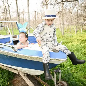 צילומי ילדים בפורים: ילד קטן בבגדי דייג וכובע יושב על חרטום סירה כחולה ביער ואוחז בחכה, לצידו פעוטה בתחפושת בת ים. צילום ילדים אמנותי ומלא דמיון בטבע מאת הצלמת שירי עציץ Shiriz Photography