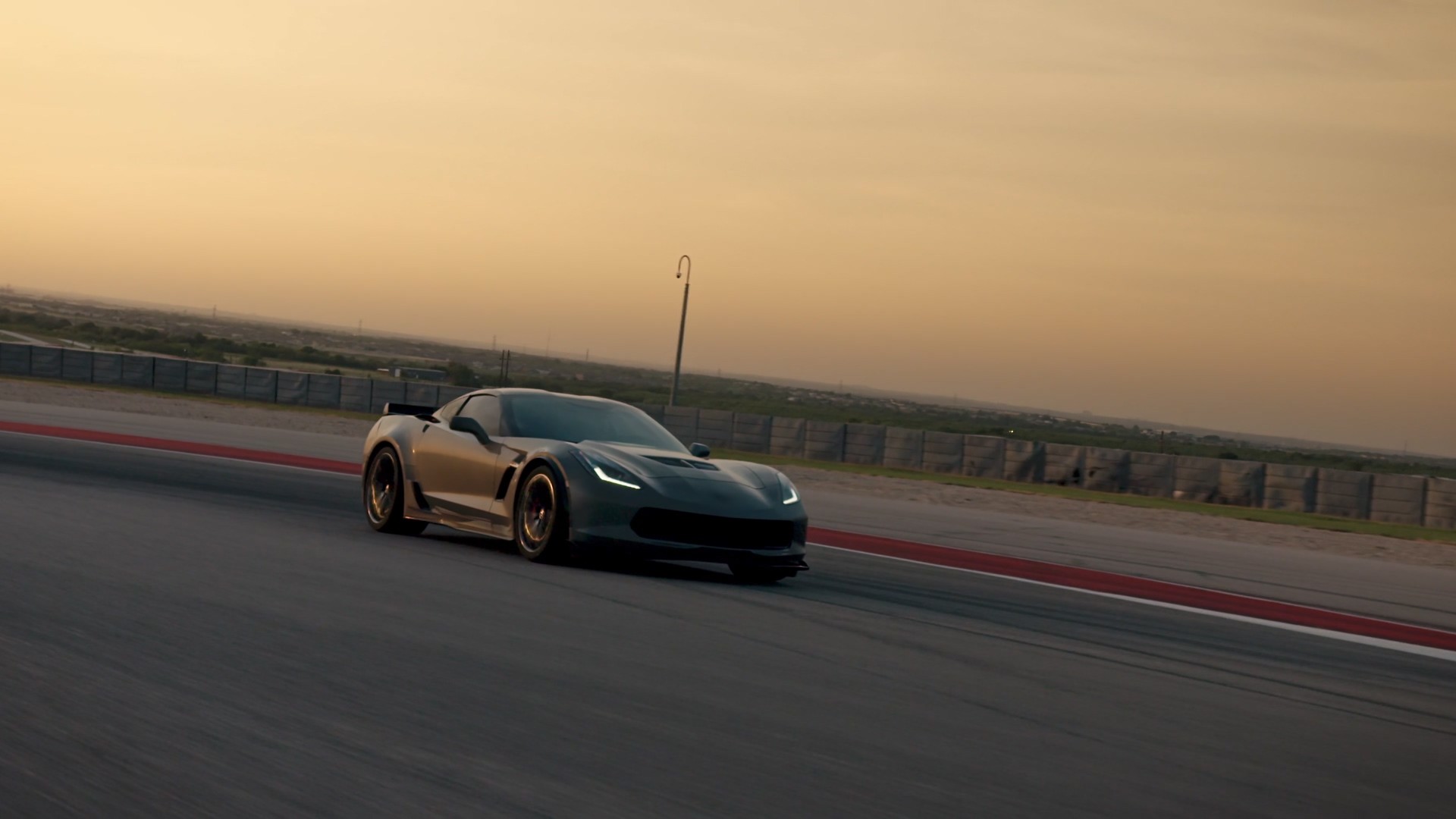 A sleek, black sports car speeds around a racetrack at sunset, highlighting its aerodynamic design and powerful performance with a scenic backdrop of an expansive sky and distant landscape.
