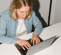 A person with blonde hair types on a laptop while seated at a table, focused on their work.