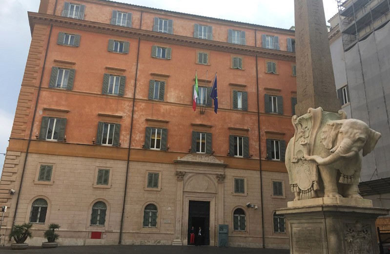 A photo of a multistore building and some roman obelisk with a statue in the middle of the square.