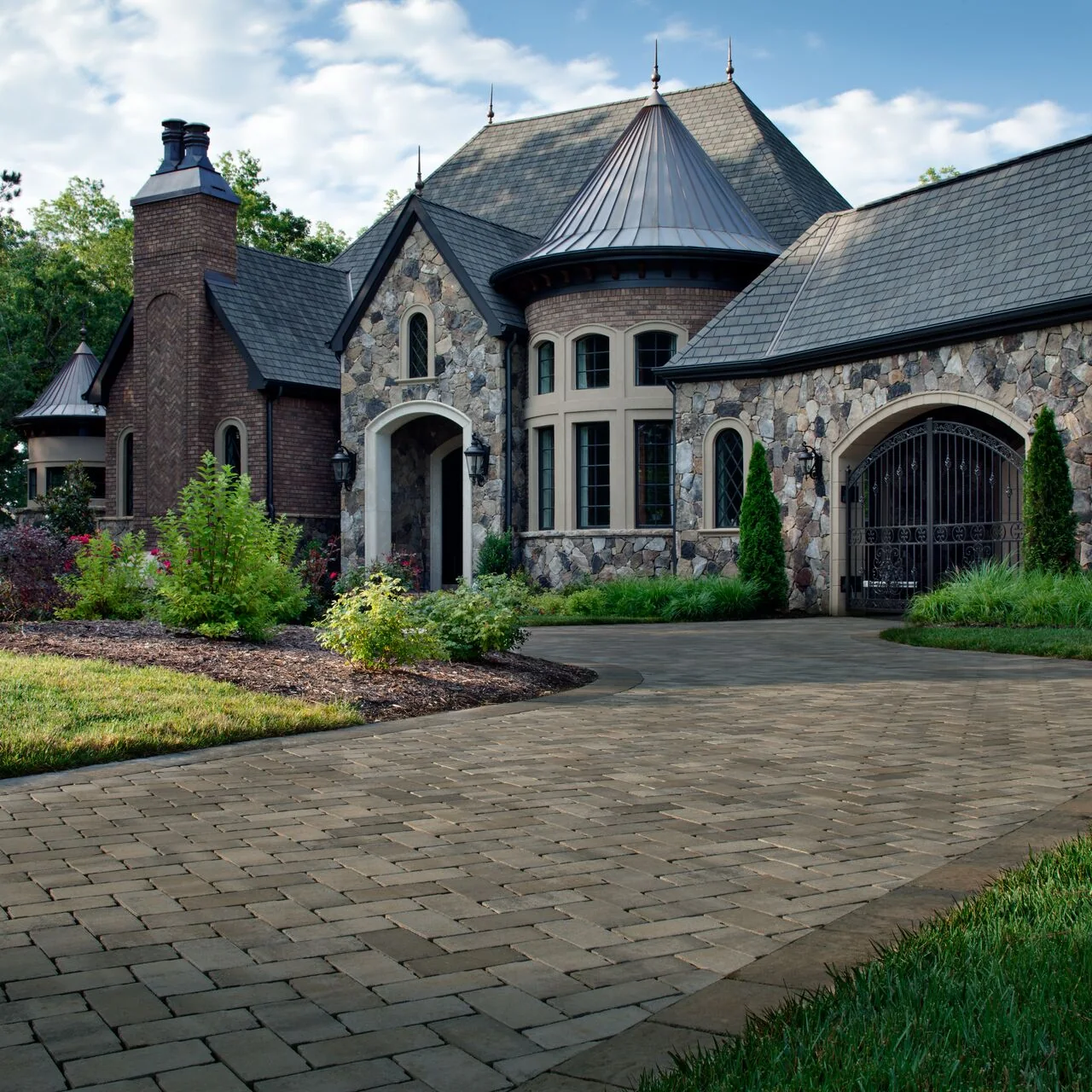 Paver arrival area in front of a stone-faced house with a turret, steep roofs and planted borders.