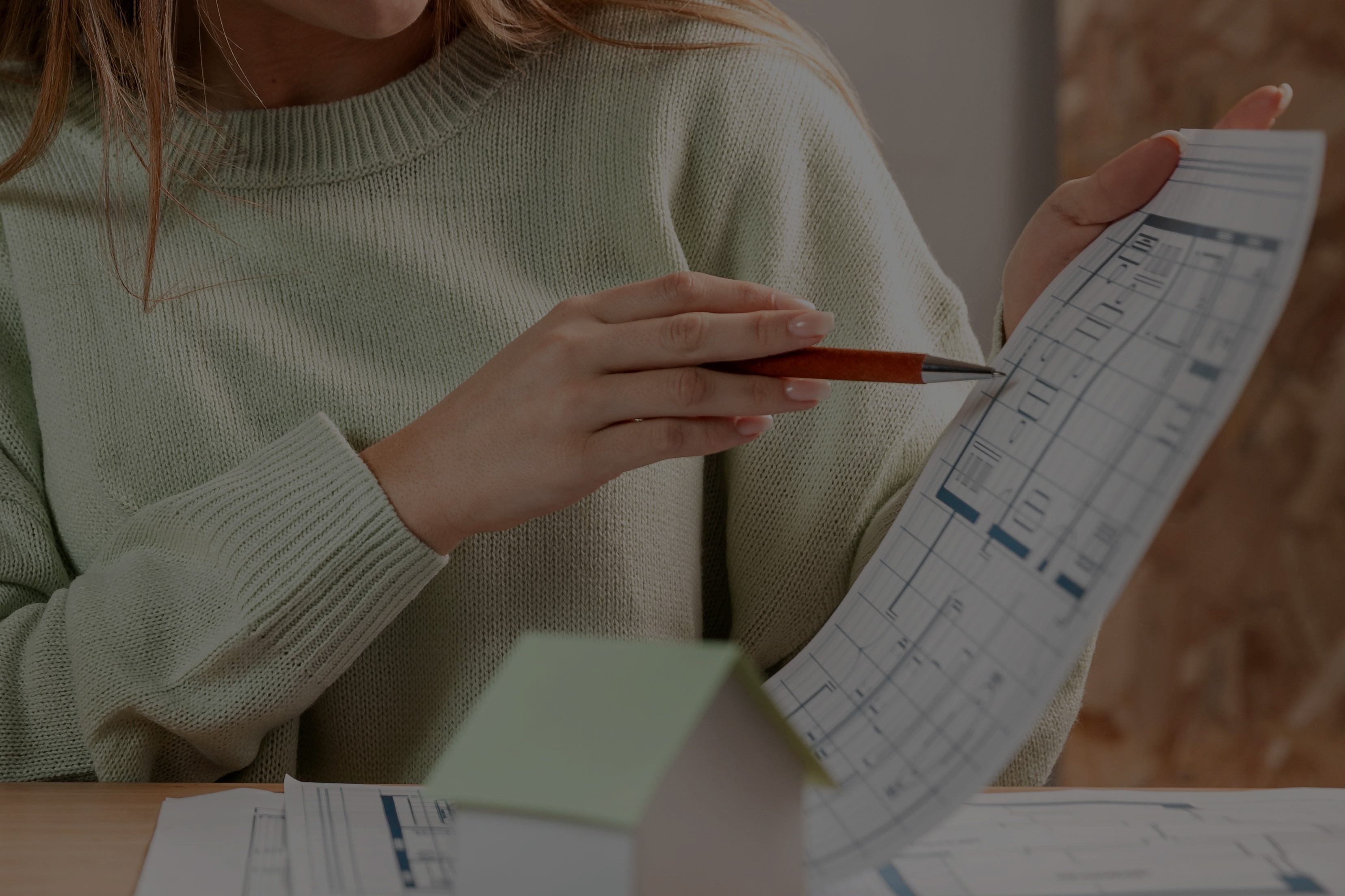 A person in a green sweater holds a pen and reviews a blueprint at a desk, with a small, paper model house in the foreground, suggesting architecture and home design concepts.