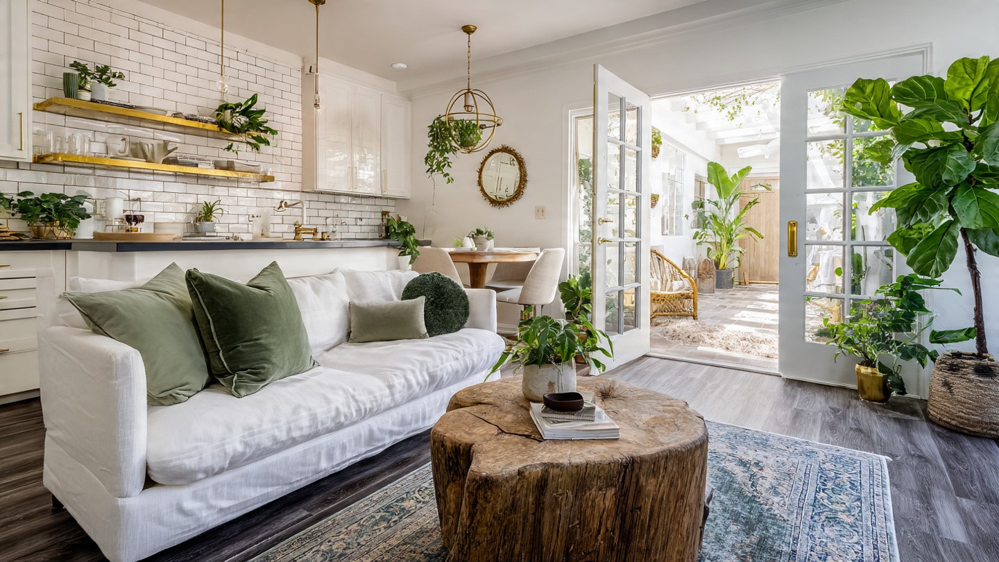 Bright living room with white sofa adorned with green cushions, a wooden coffee table, abundant plants, and open French doors to a sunlit patio.
