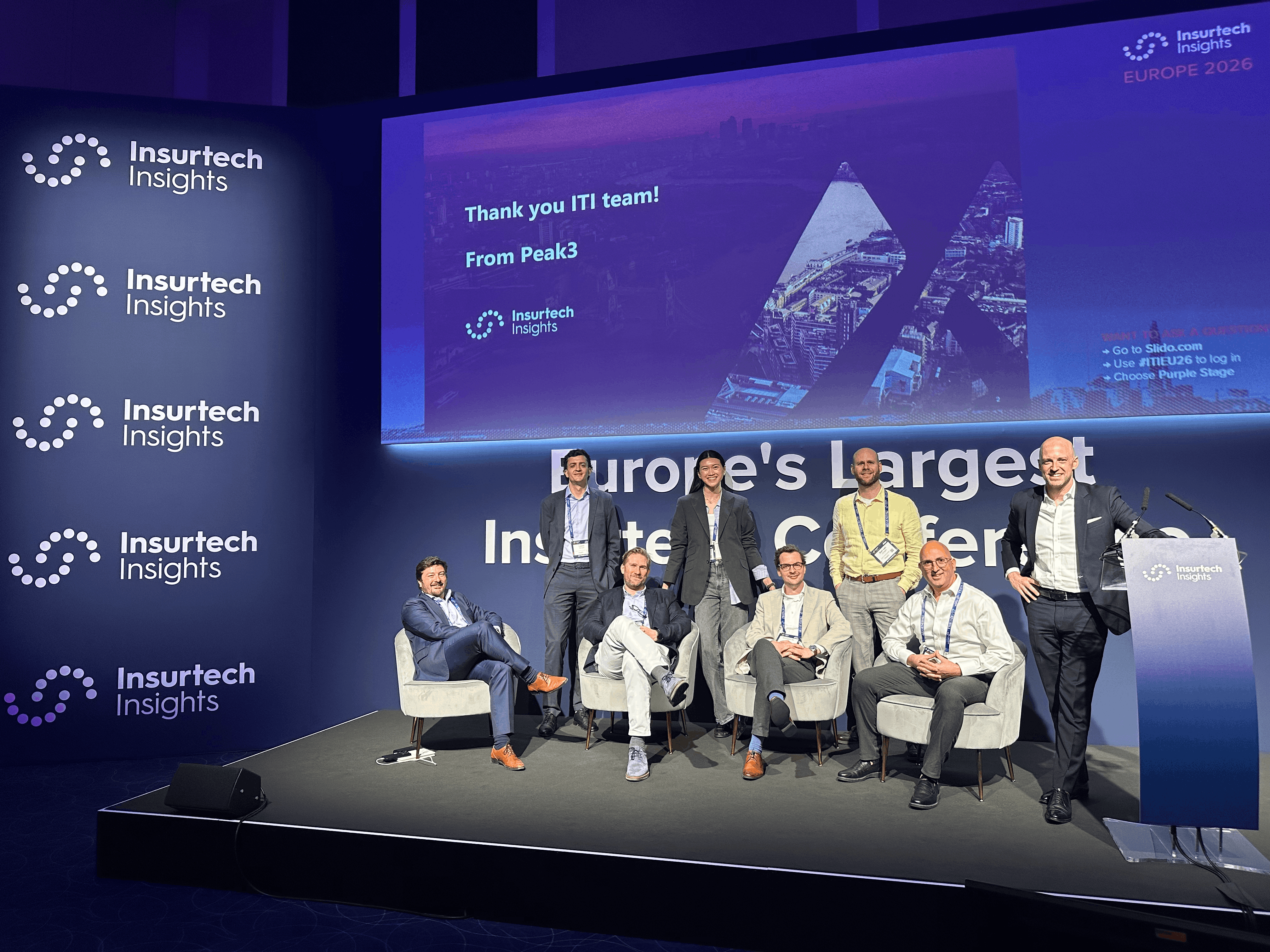 Panel discussion with five speakers on stage at an event, with a blue backdrop displaying event branding.