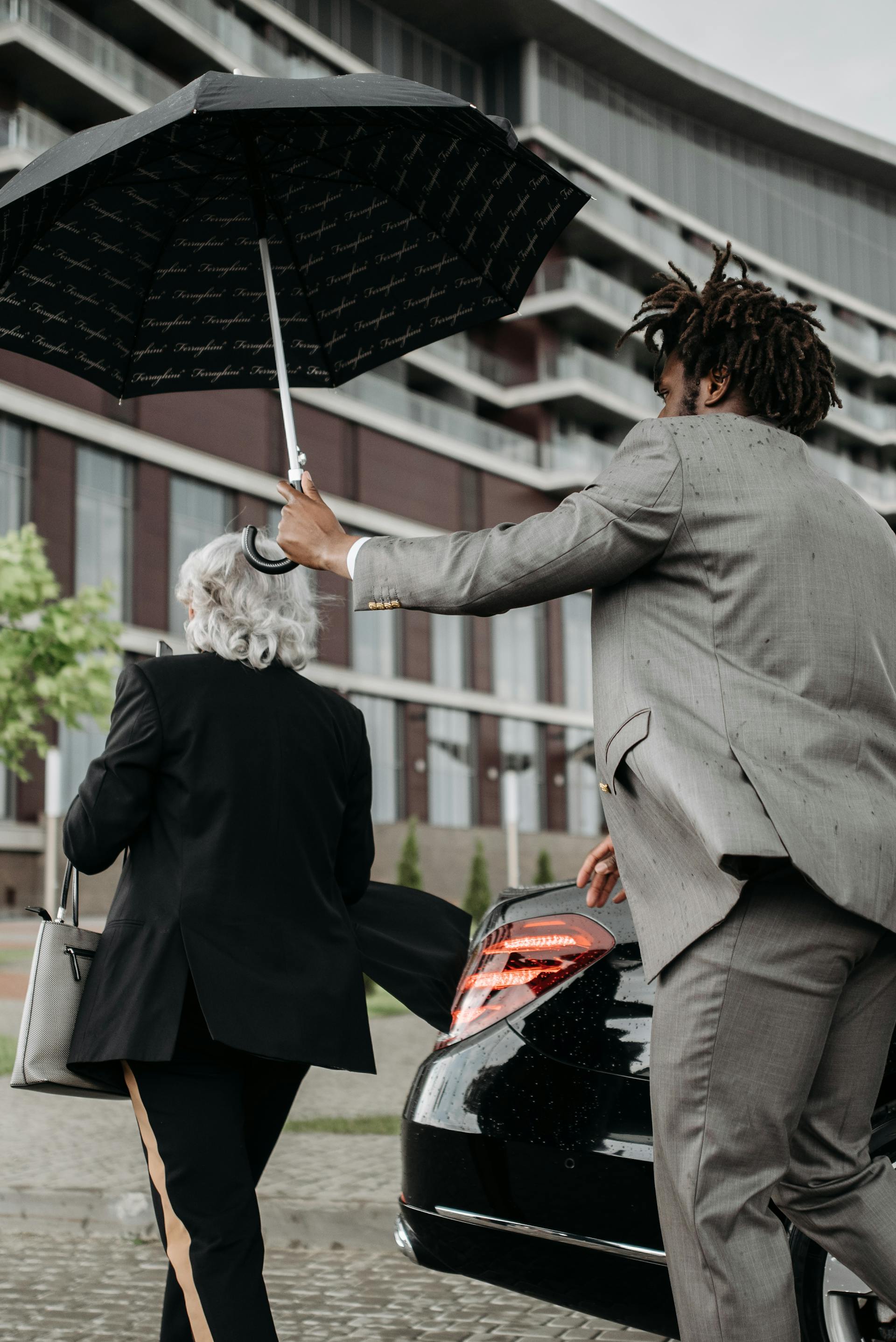 Luxury chauffeur assisting a client with an umbrella while approaching a black sedan, showcasing premium transportation service.