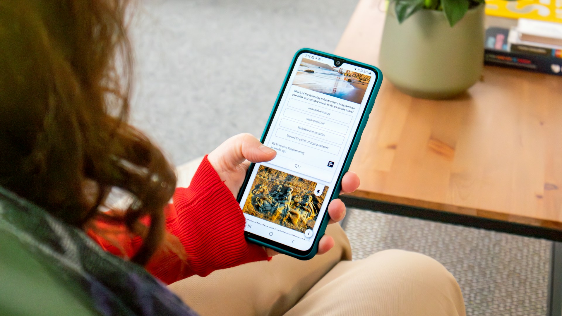 A woman is sitting on a couch, holding a smartphone with a green case and scrolling through a feed that displays a combination of text and images.