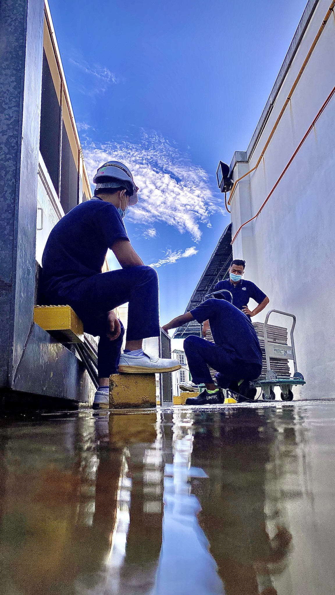 A young person with curly hair smiles at the camera while sitting in front of a laptop against a dark background.