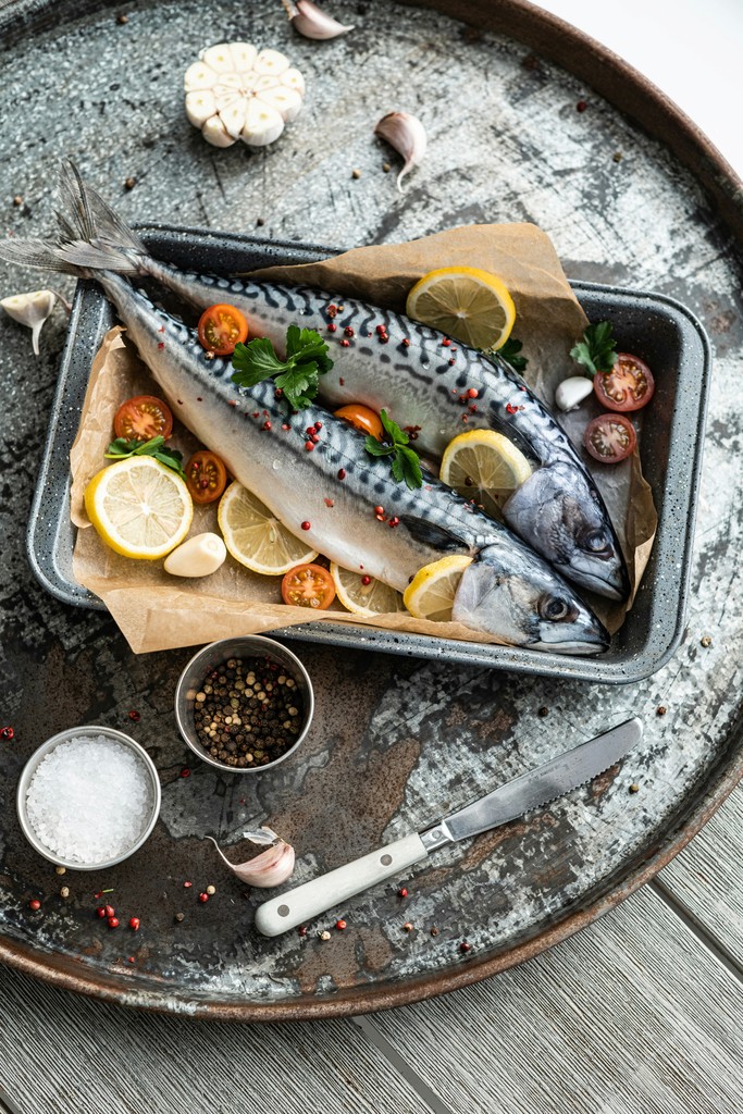 Fresh mackerel prepared for baking, arranged in a metal tray with lemon slices, cherry tomatoes, garlic cloves, parsley, and peppercorns, set on a rustic metal surface with salt and a knife nearby.