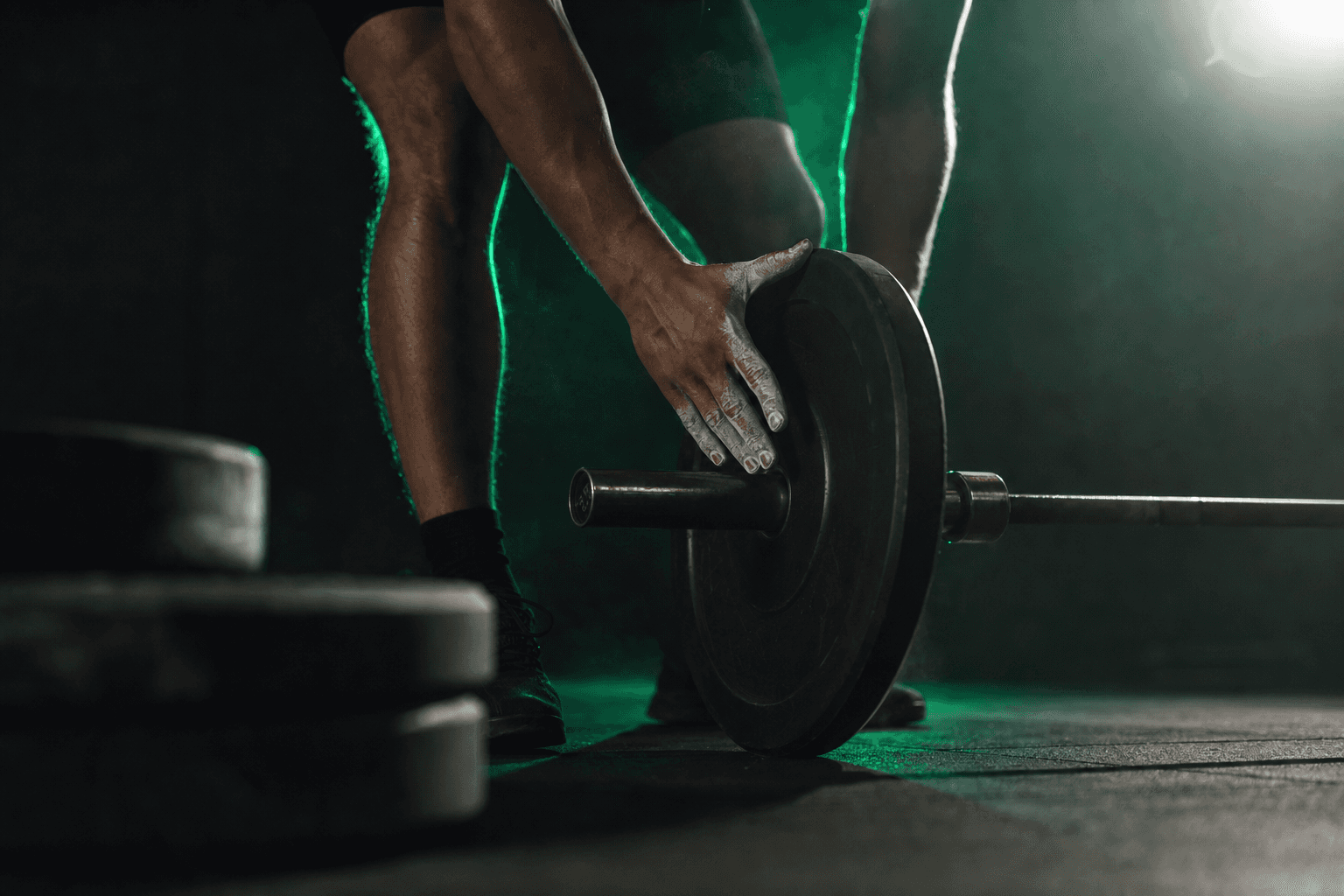 A lifter's chalked hands loading fewer, lighter plates onto a barbell, with heavier plates set aside in shadow, lit by a green accent