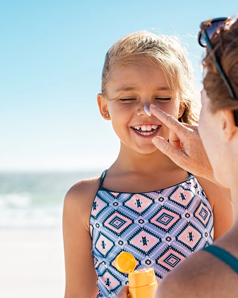 A mother applying sunscreen to her young daughter's nose on a sunny beach