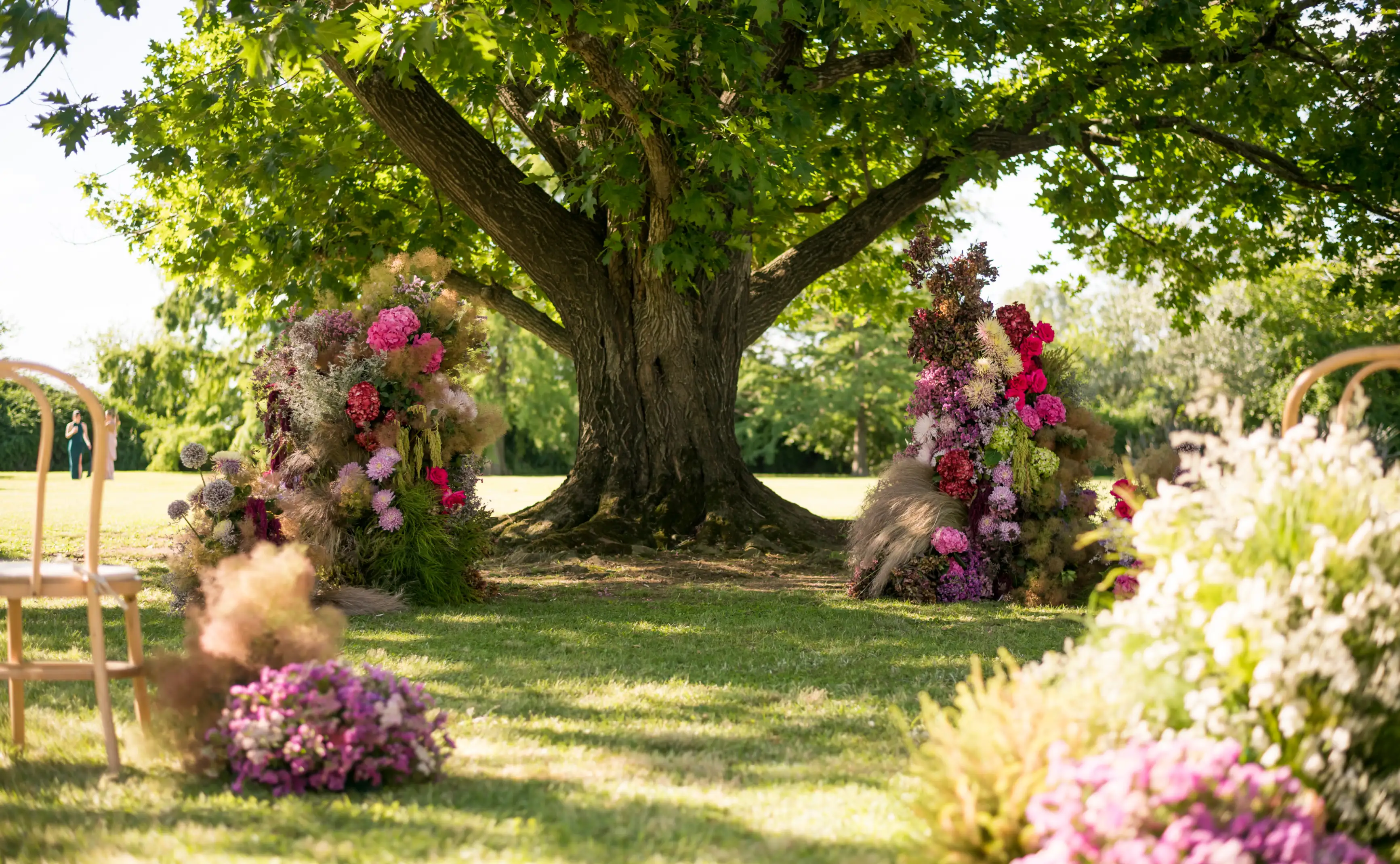 Ceremonia al aire libre bajo un gran árbol, decorada con estructuras florales en tonos rosados, lilas y verdes.