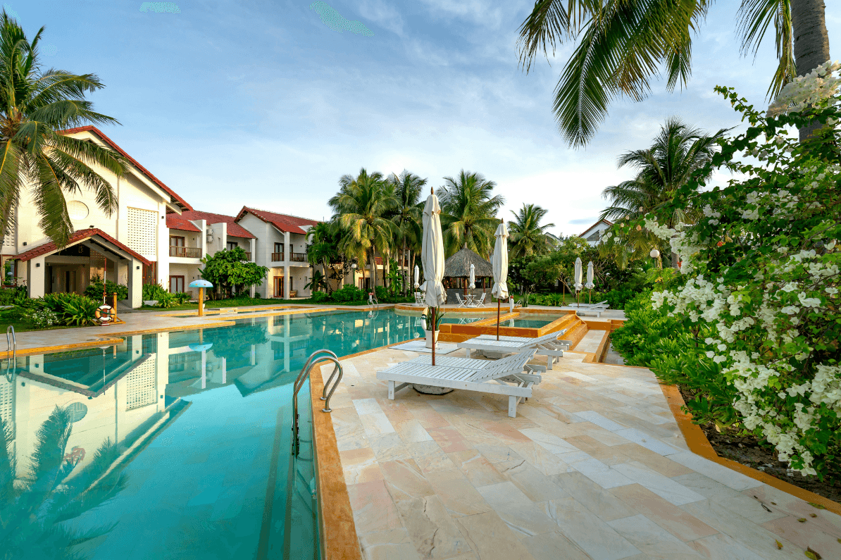White resort bungalow with wide porch, potted plants, landscaped lawn, surrounded by palm trees.
