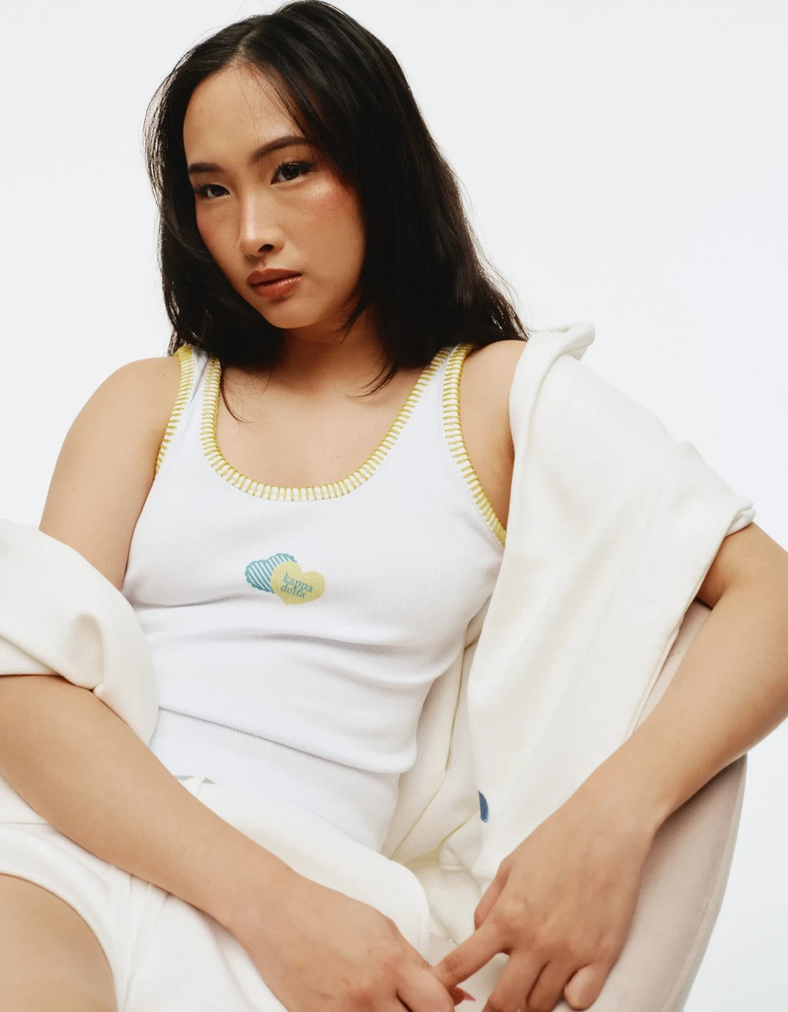 Studio portrait of a model wearing a white Kappa Delta tank with yellow trim and matching white shorts, with a cream hoodie draped off the shoulders.