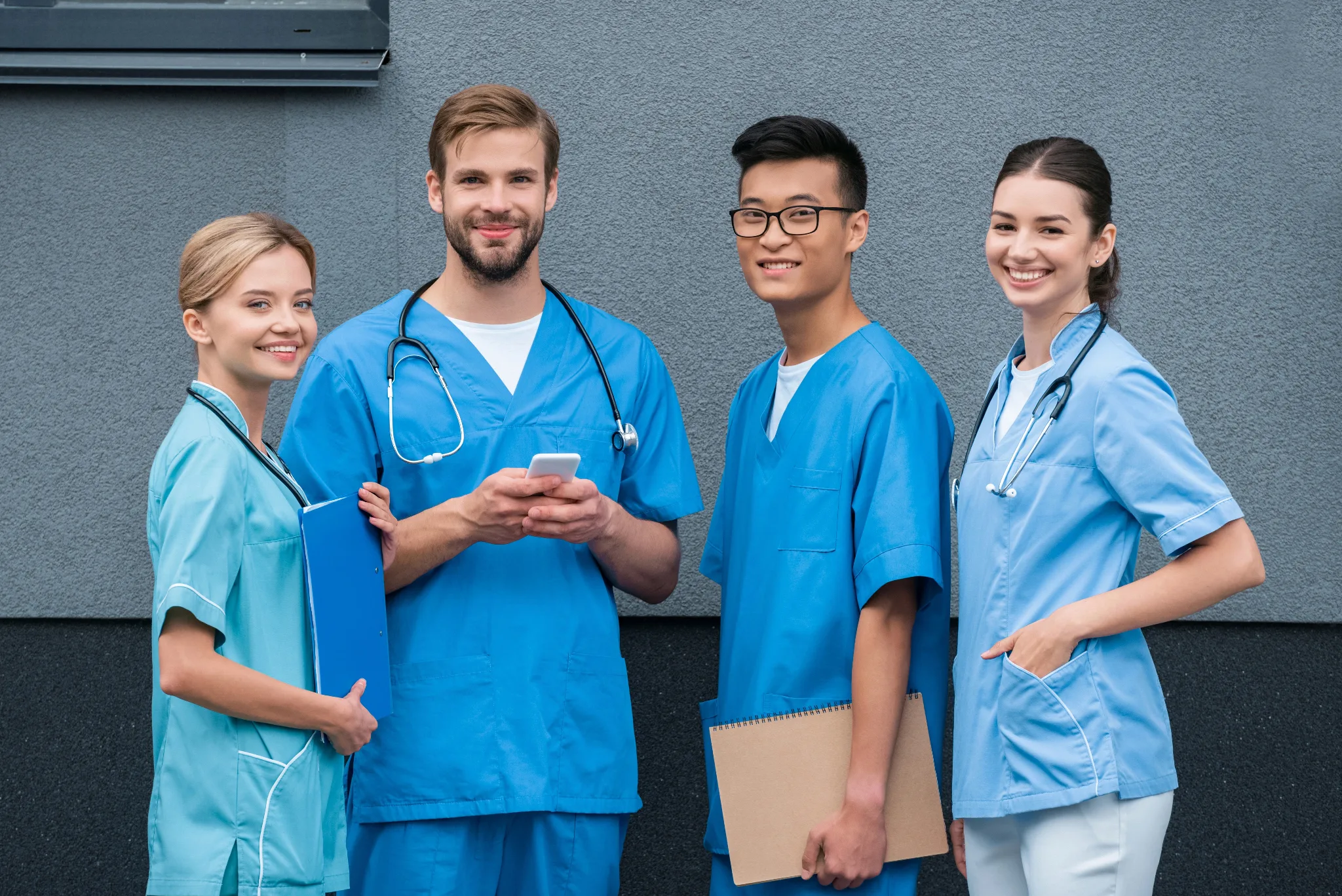 Four smiling healthcare professionals in blue medical scrubs stand together against a gray wall, with one holding a smartphone and another carrying a clipboard.