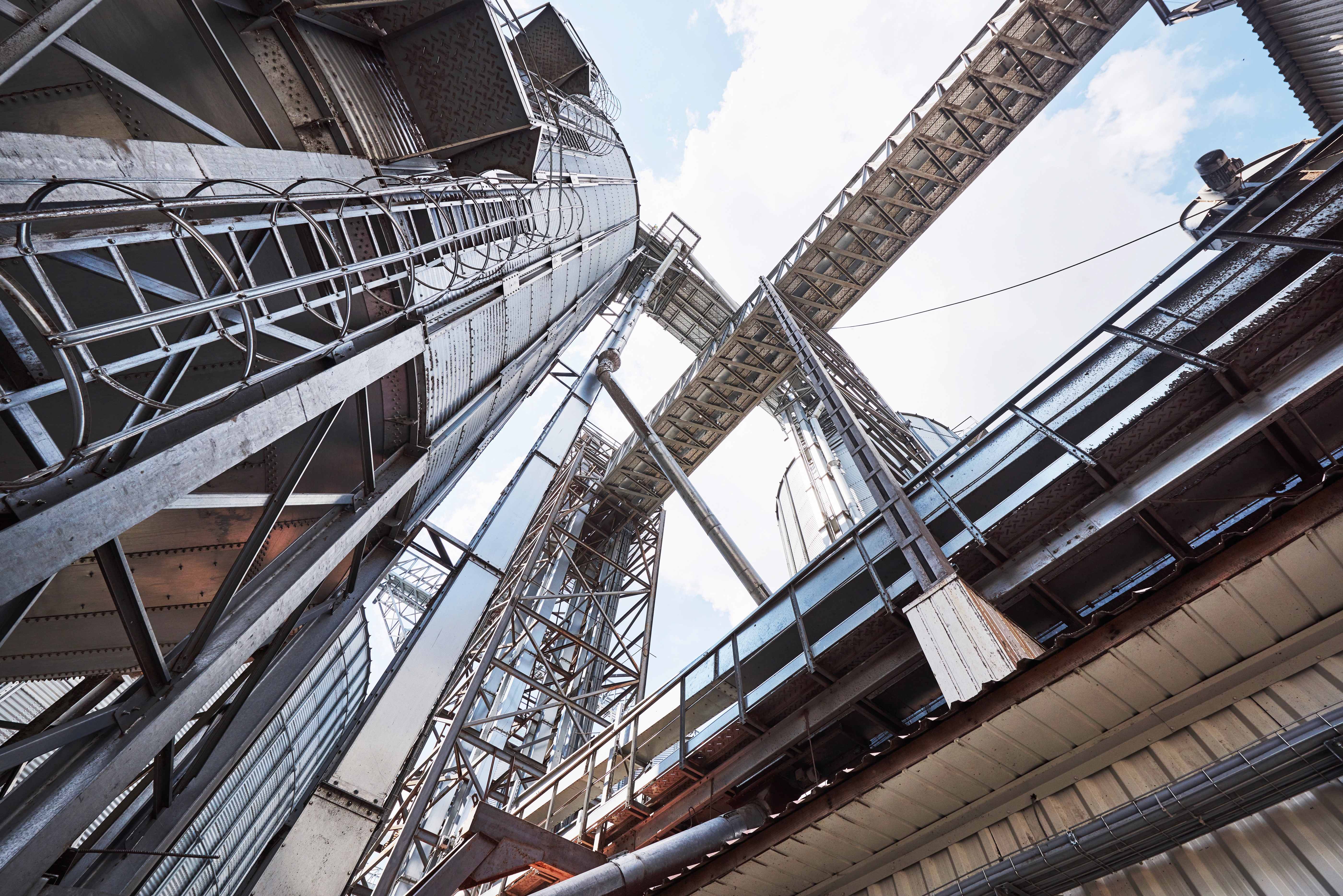 Upward view of industrial silos and metal structures against a blue sky. The steel beams and walkways create a complex, geometric pattern, conveying a sense of scale and industry.