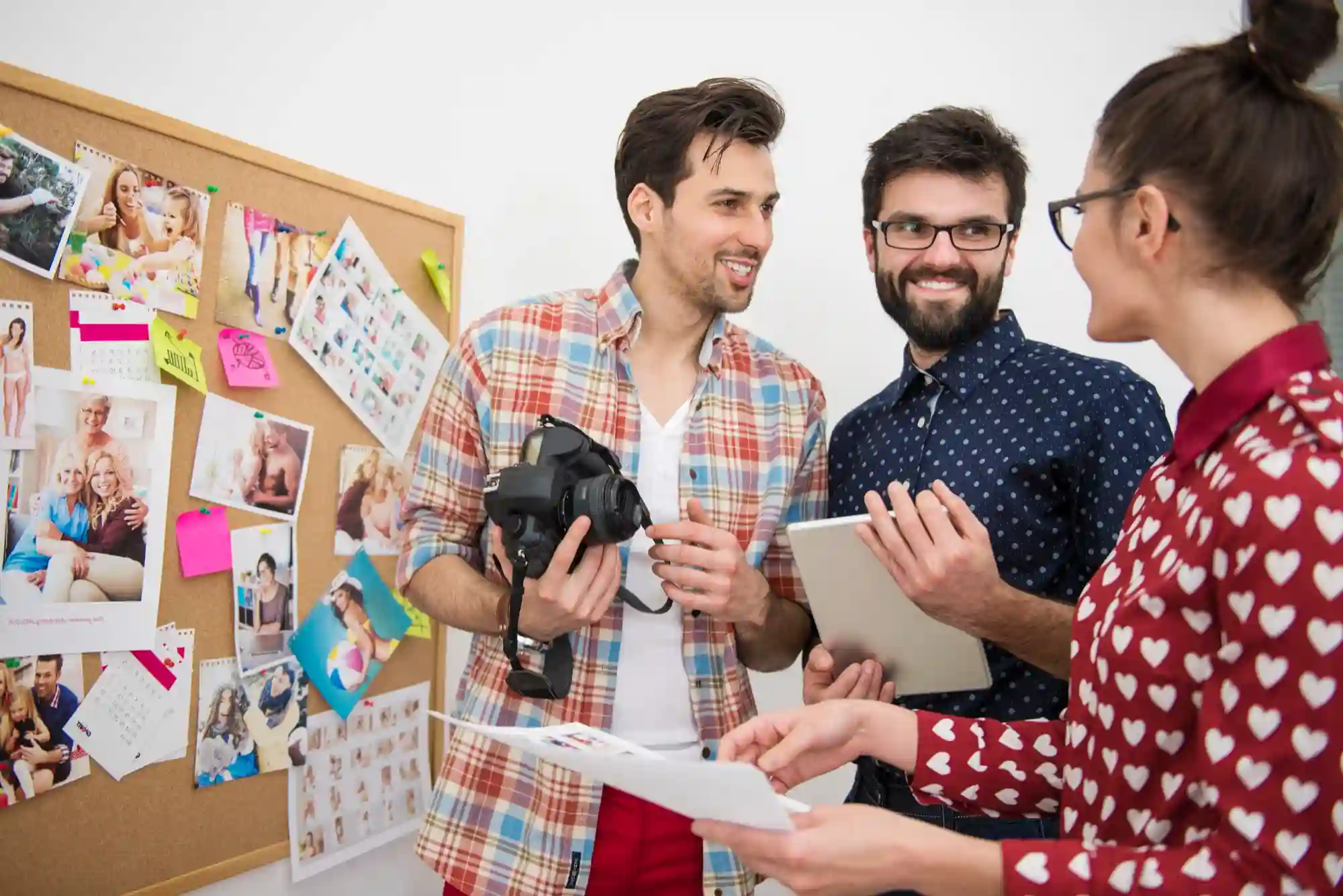 A diverse creative team collaborates in an office, reviewing photos on a corkboard and discussing projects with a camera.