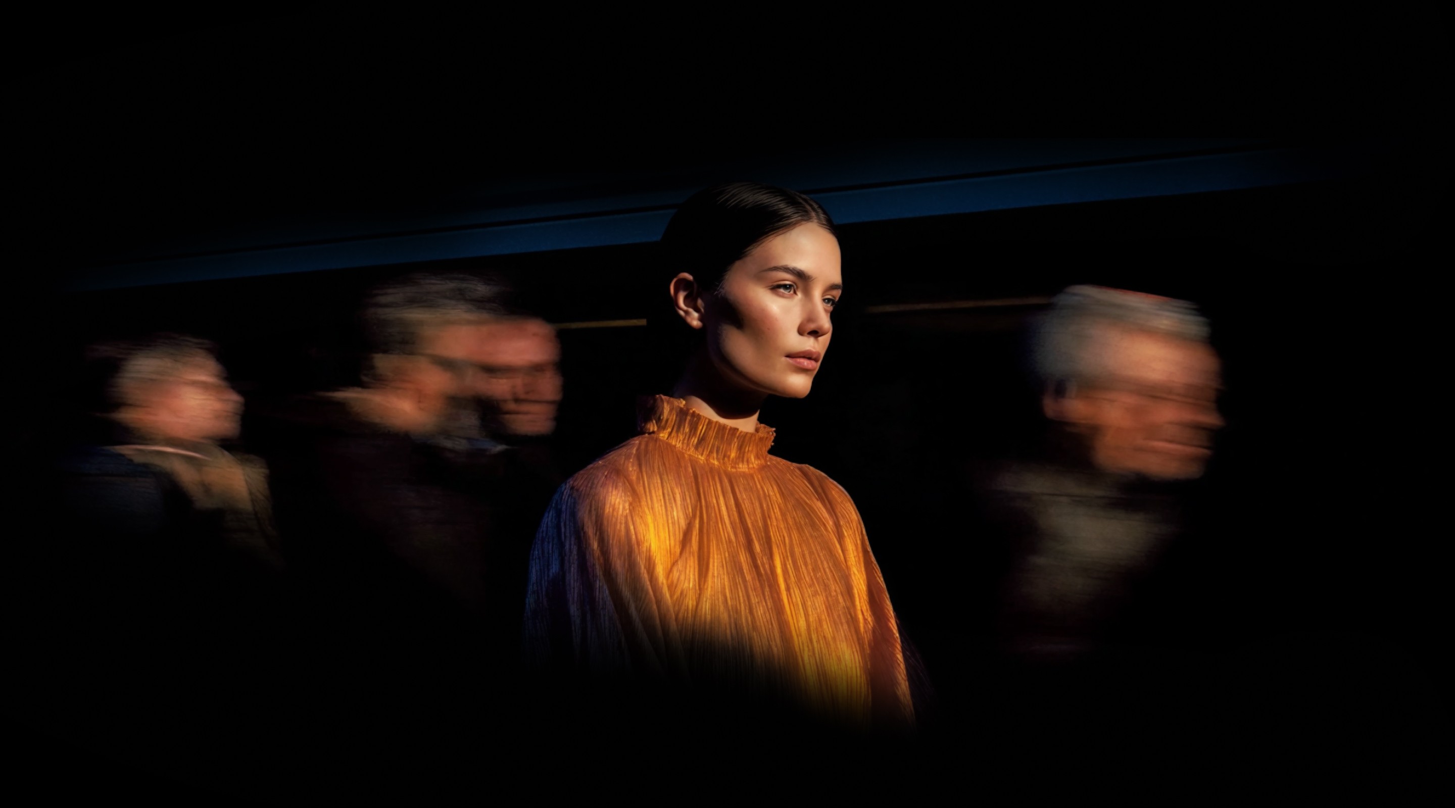 Portrait of a woman in warm lighting standing still while blurred people move around her in a dark environment.