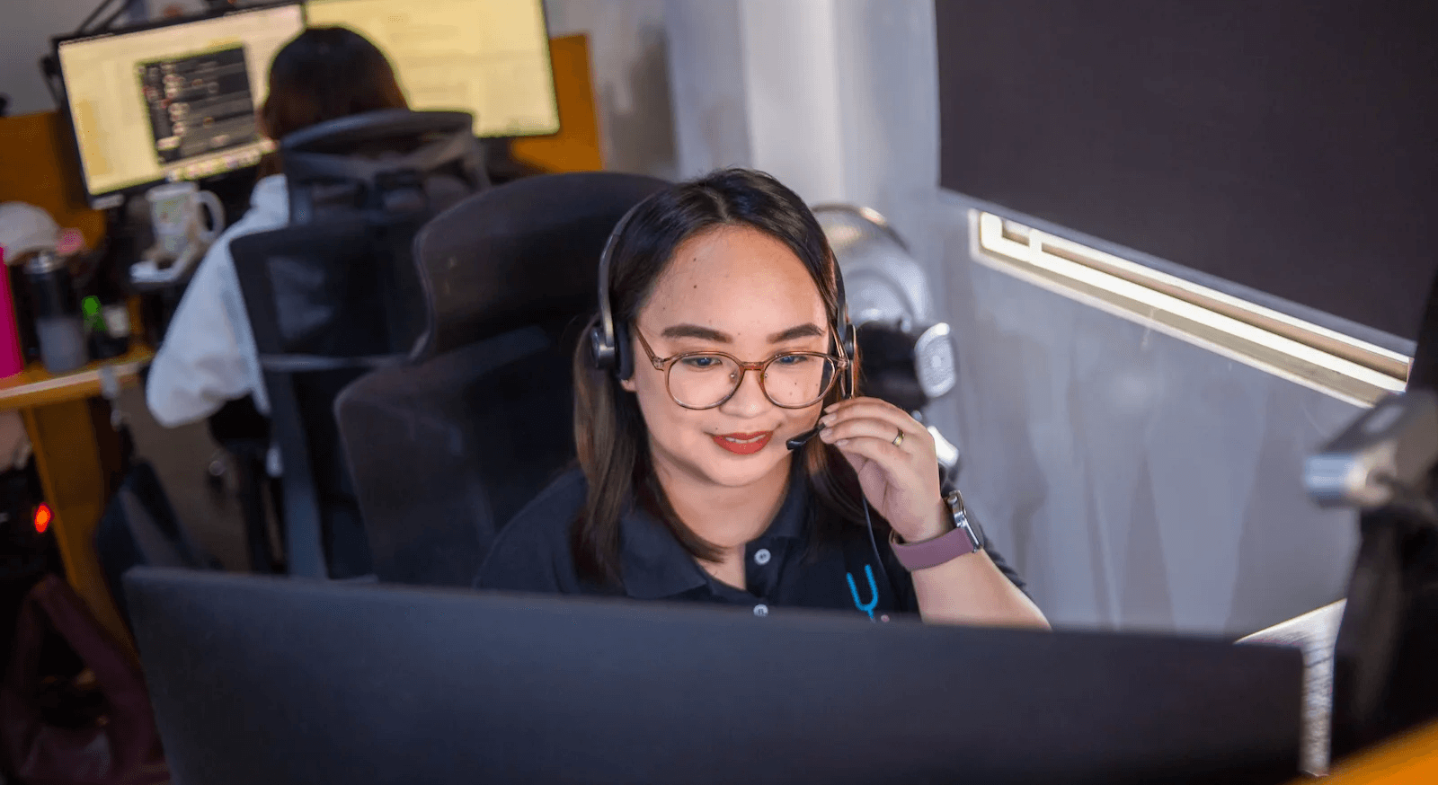 A woman wears glasses and smiles while working at a computer in a dimly lit workspace.