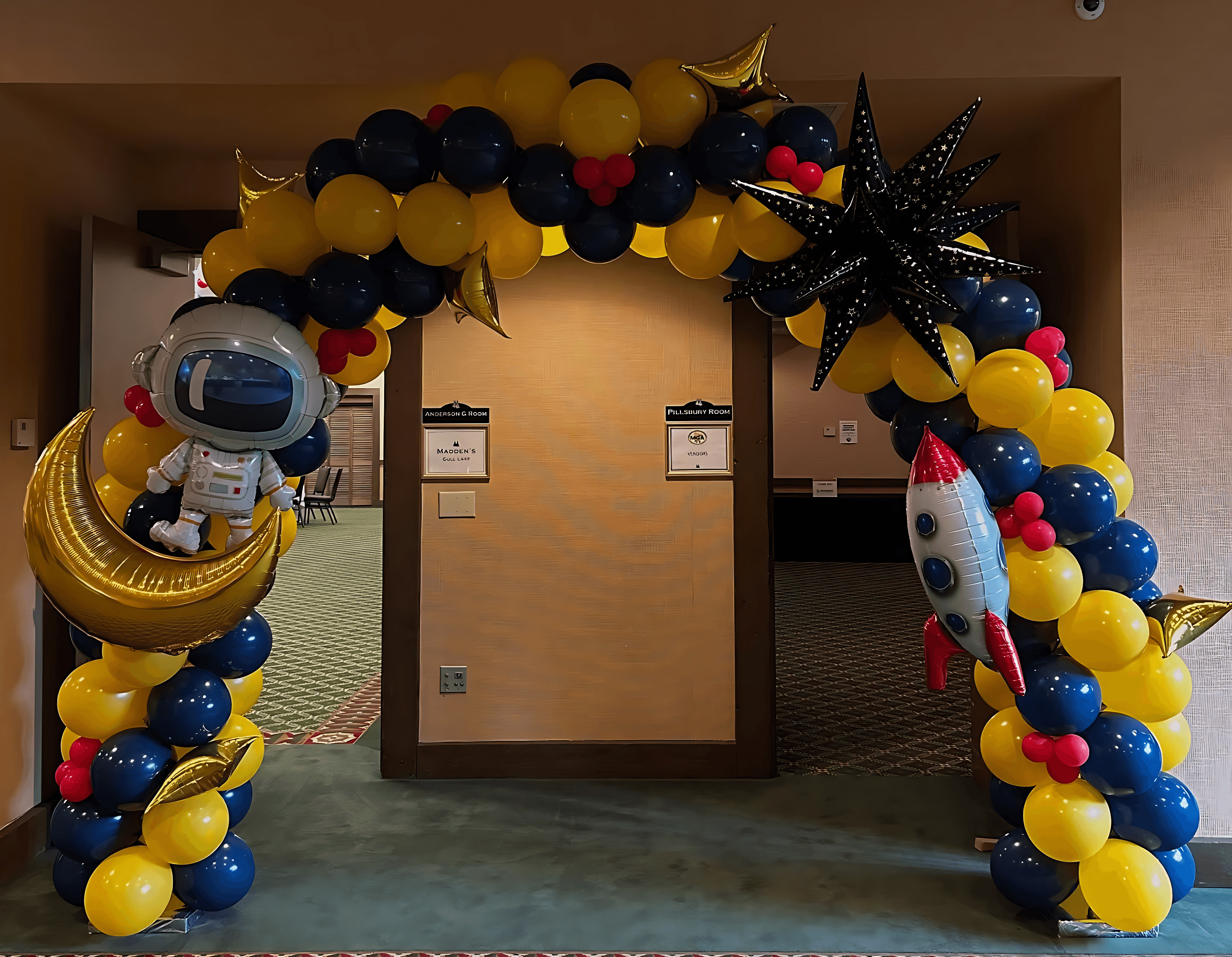 A festive balloon arch featuring bright yellow and blue balloons, adorned with a cartoon astronaut on a crescent moon and a small red and white rocket, creates a lively entranceway, ideal for a space-themed event.