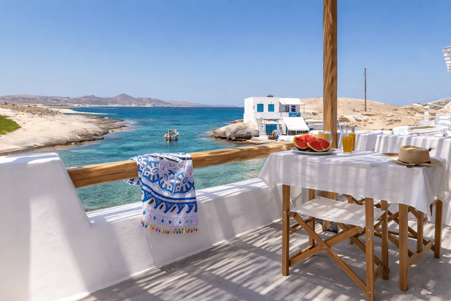 Seaside balcony with table set for lunch, overlooking blue ocean and white buildings under a sunny sky.