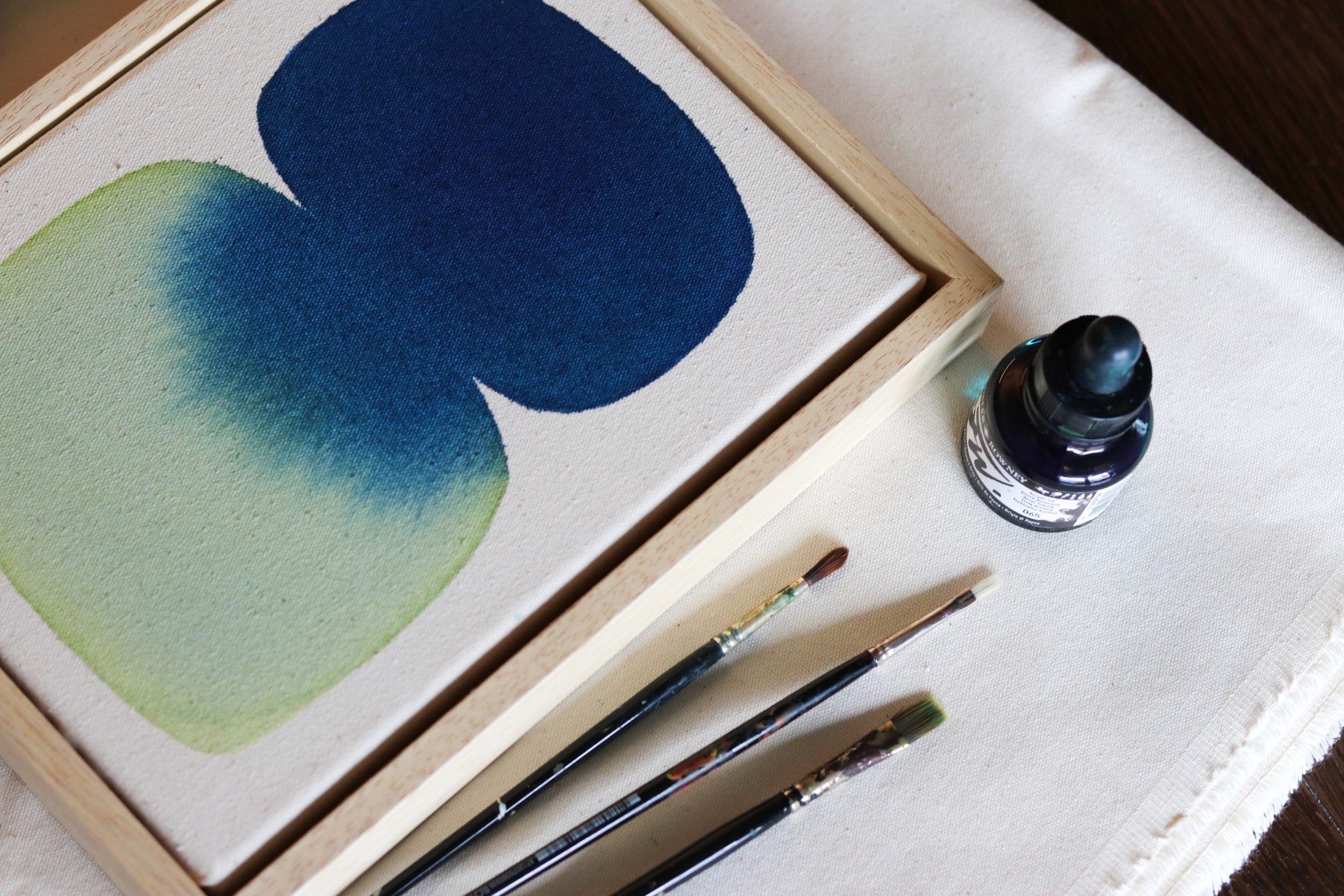 An artist's workspace featuring a watercolor painting with blue and green swatches, paintbrushes, and ink.