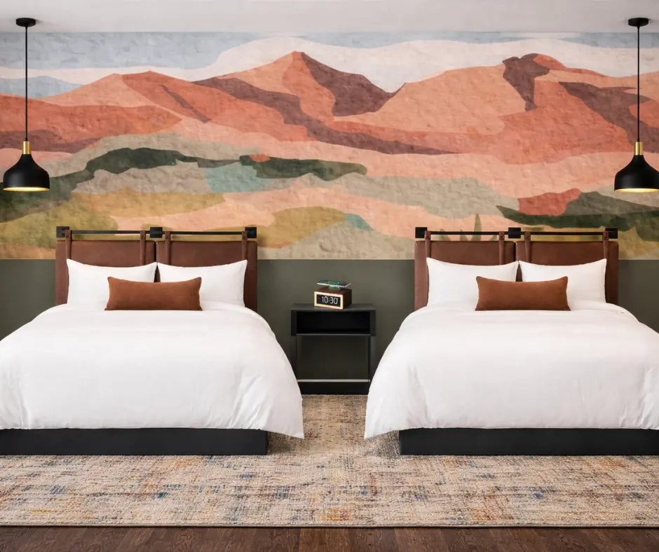 A modern hotel room featuring two beds with white linens, set against a desert landscape mural.