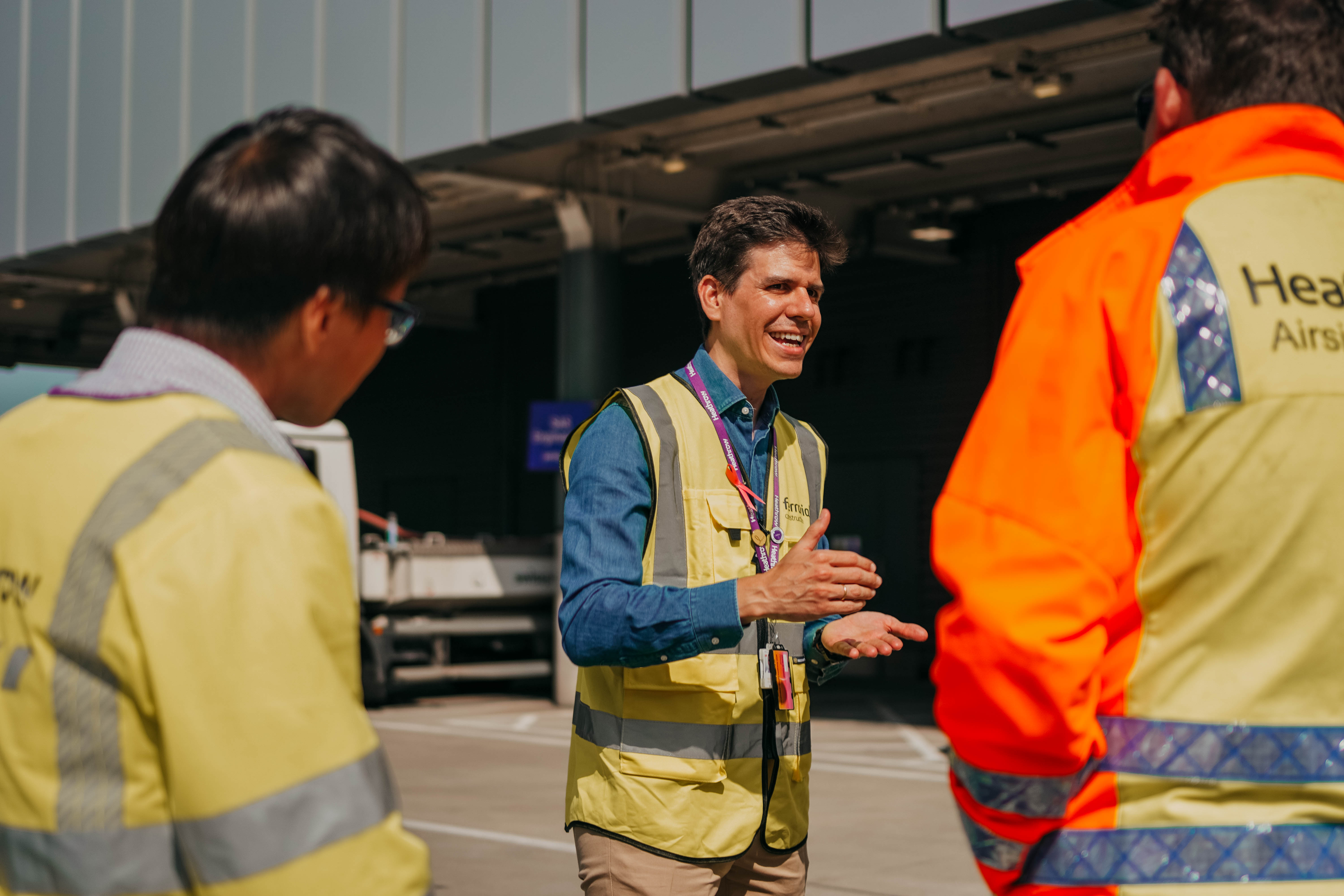 A man in a yellow high-visibility vest over a denim shirt smiles and gestures as he talks with two other airport workers on a sunny tarmac.