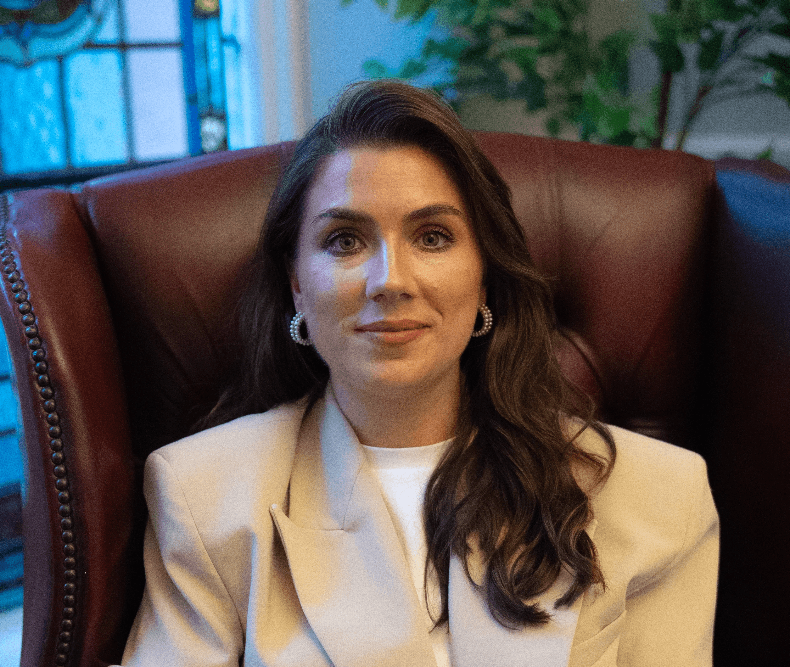 A person with curly hair and glasses smiles at the camera, seated in a cozy workspace with warm lighting.