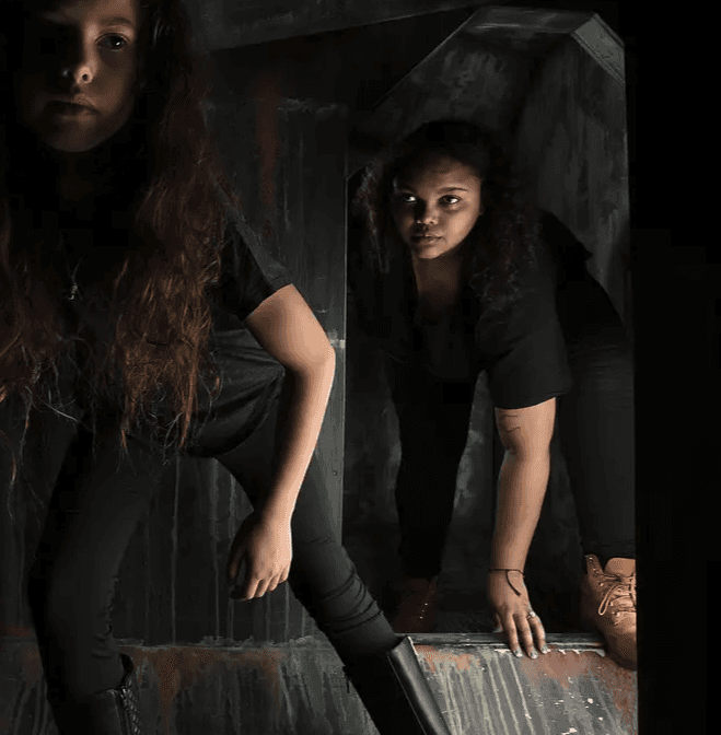 A young person with curly hair smiles at the camera while sitting in front of a laptop against a dark background.