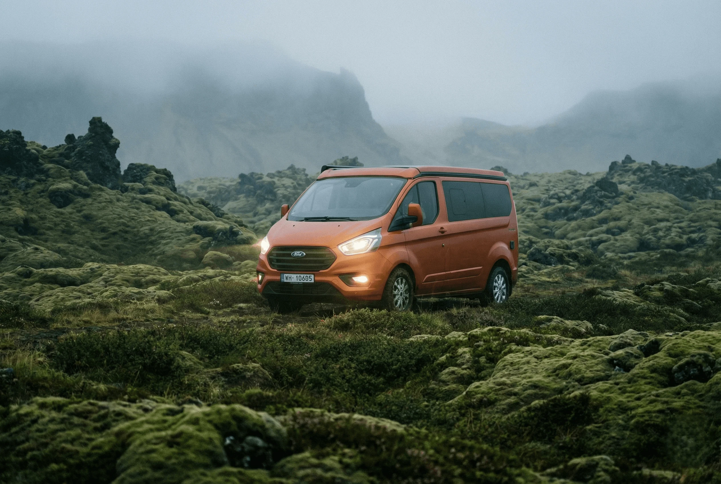 An orange camper van parked in a green, moss-covered lava field with fog in the distance.
