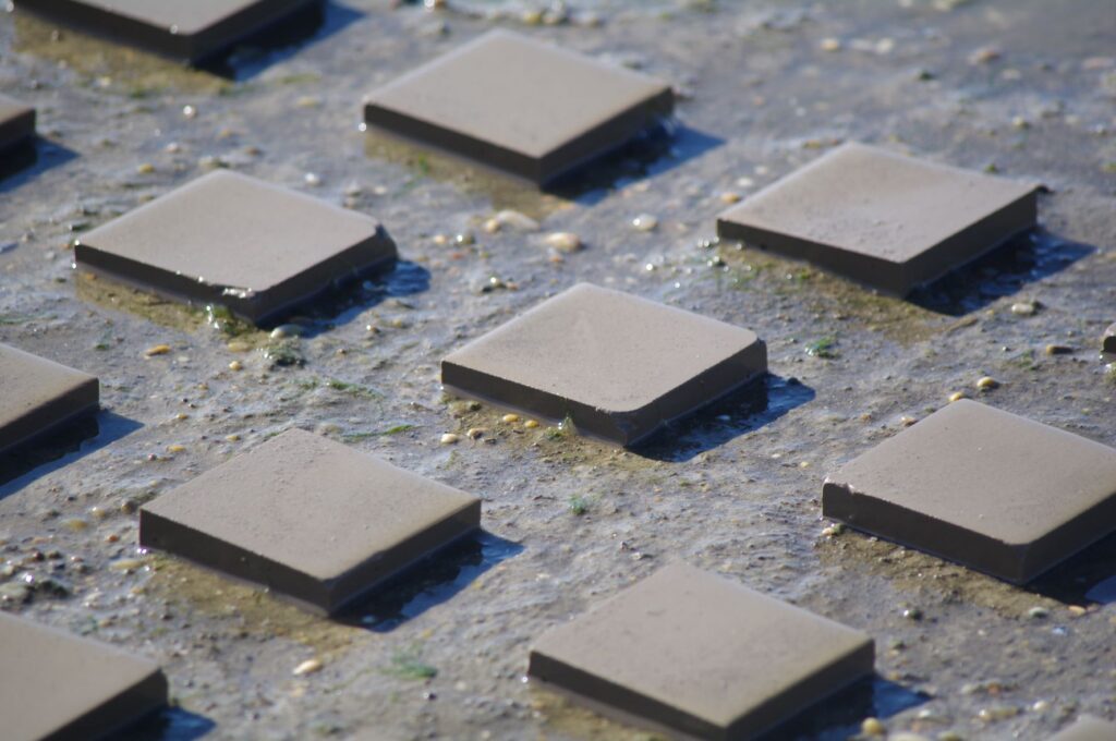 Dalles carrées en céramique gris anthracite disposées en quinconce sur du sable mouillé avec coquillages, testées en conditions d'immersion extérieure
