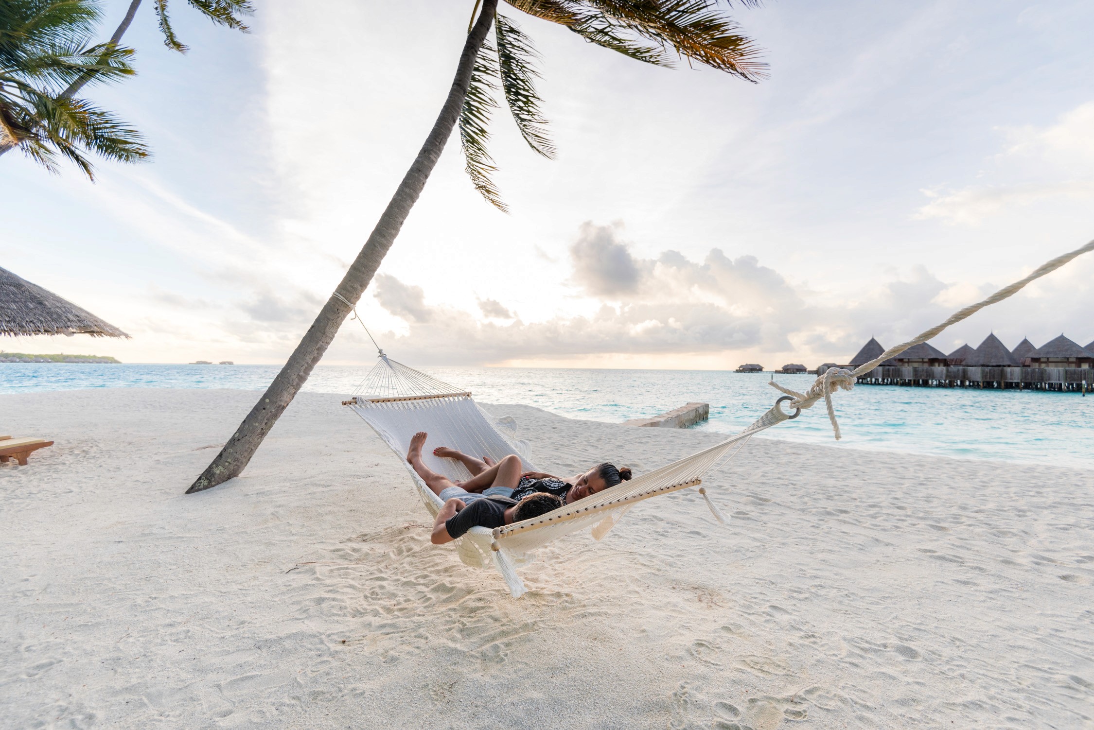 A person lies in a white hammock strung between two palm trees on a white sand beach with turquoise water and huts.
